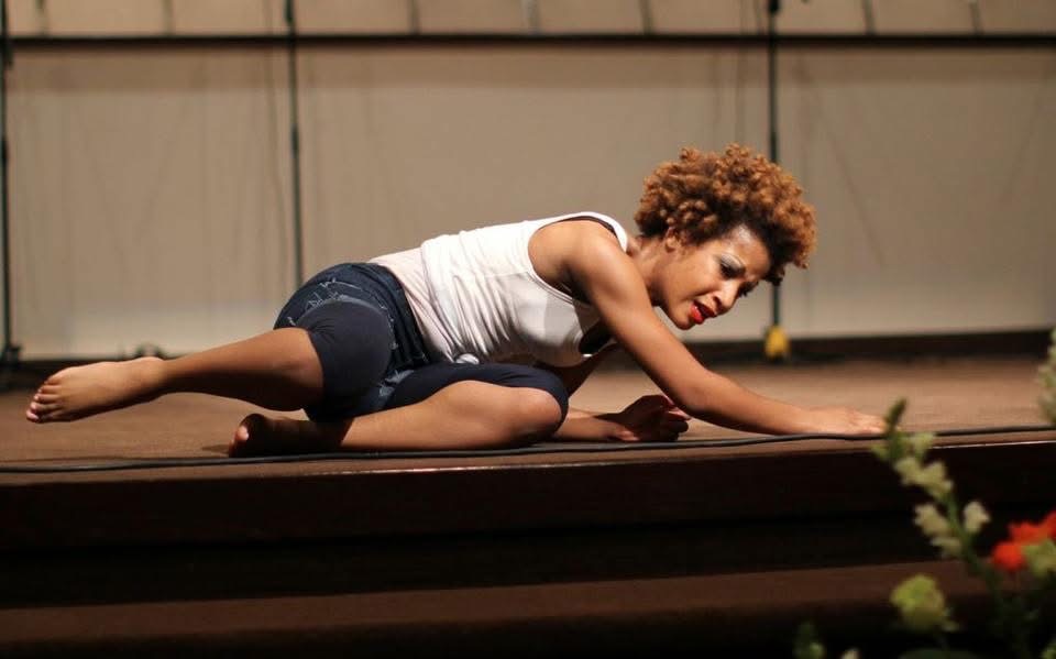 Child with curly hair lying on stage, supporting with one arm, in a white tank top and dark shorts.
