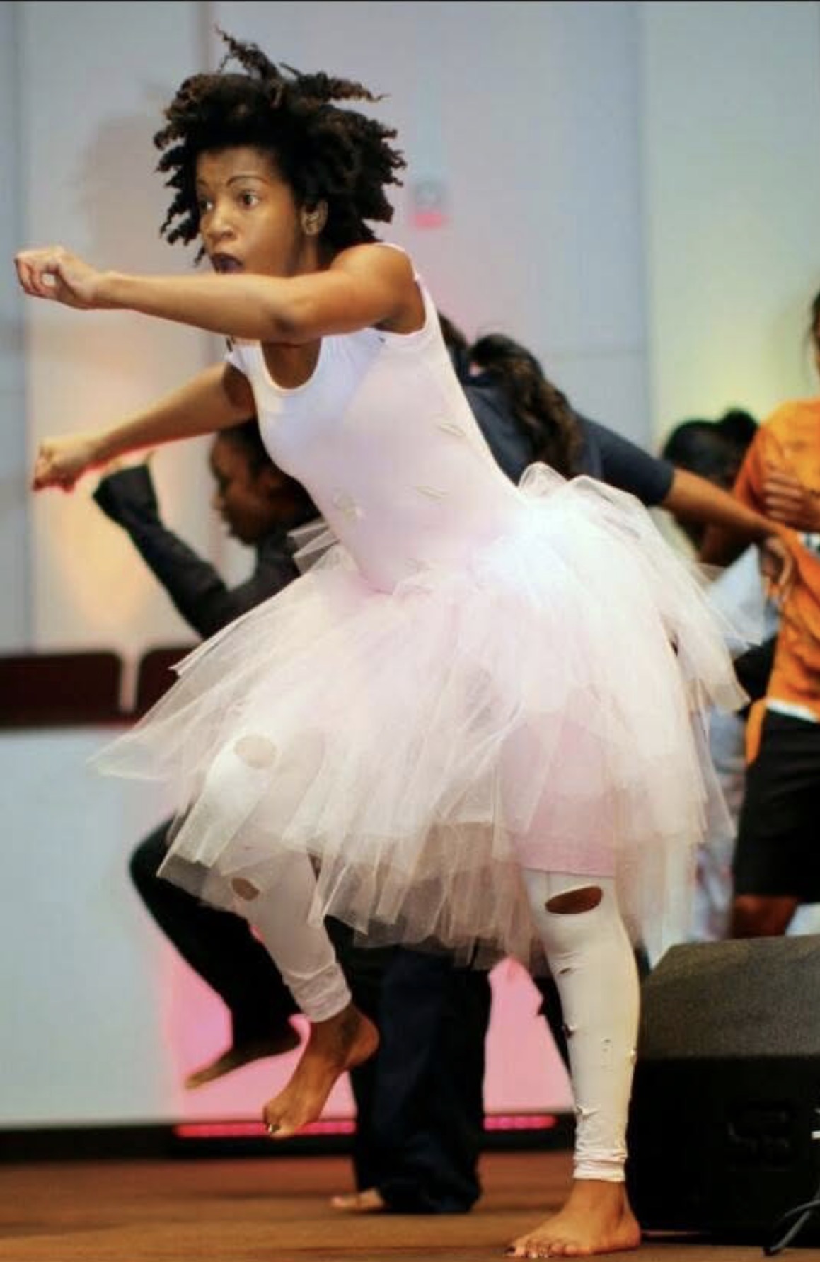 Young girl in a white tutu dance costume performs on stage with other children in background.