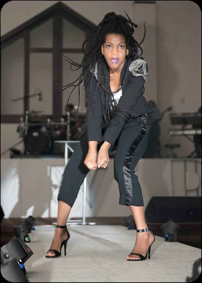 Woman with dreadlocks in black outfit and high heels performing on stage, with musical instruments in background.