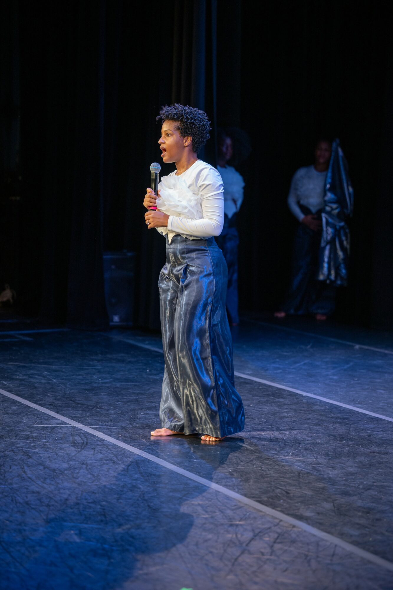 Person standing on stage holding a microphone, wearing a white top and shiny pants, with a dark background.