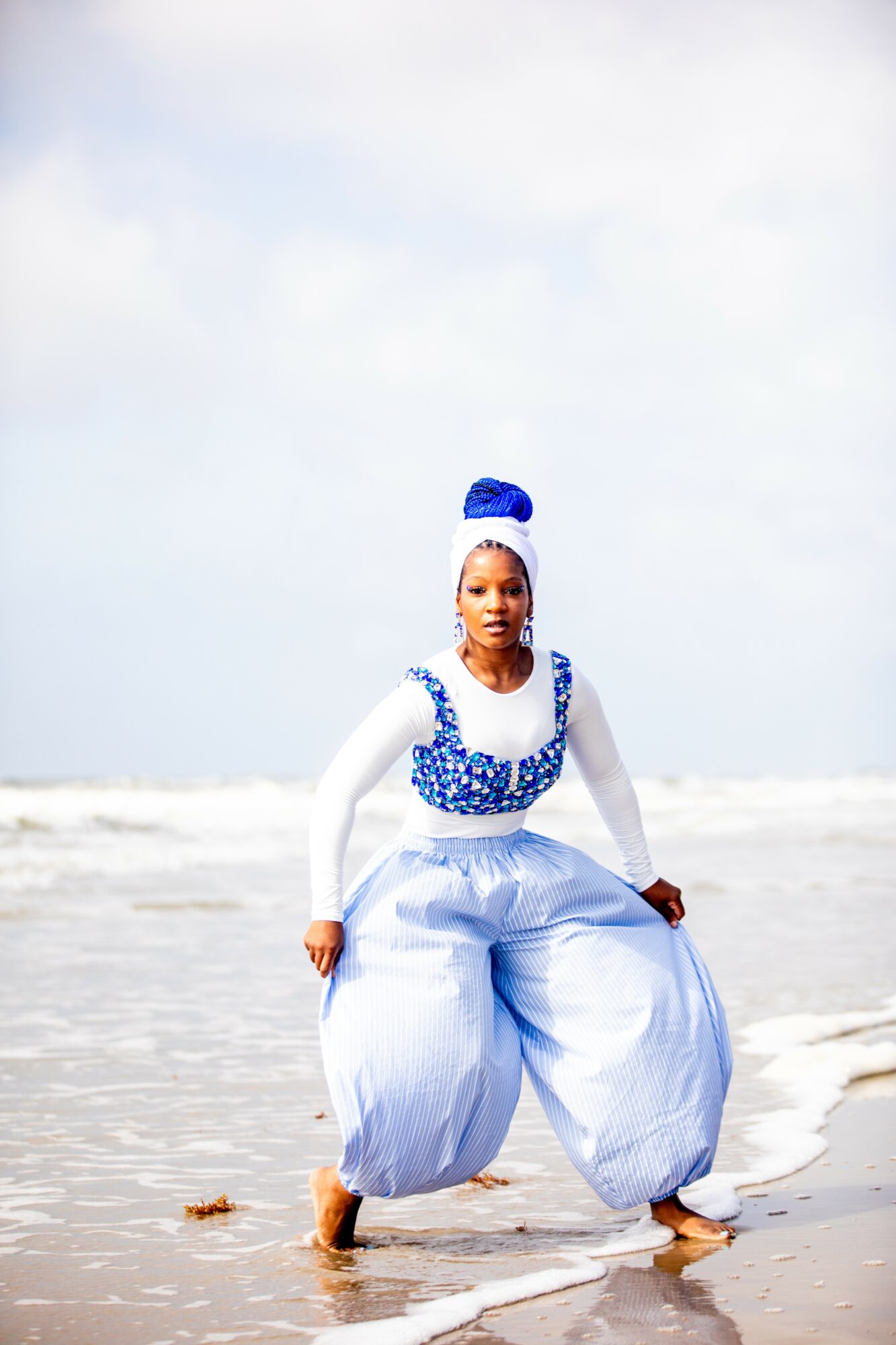 Woman in blue and white outfit standing on beach with ocean and sky in background.