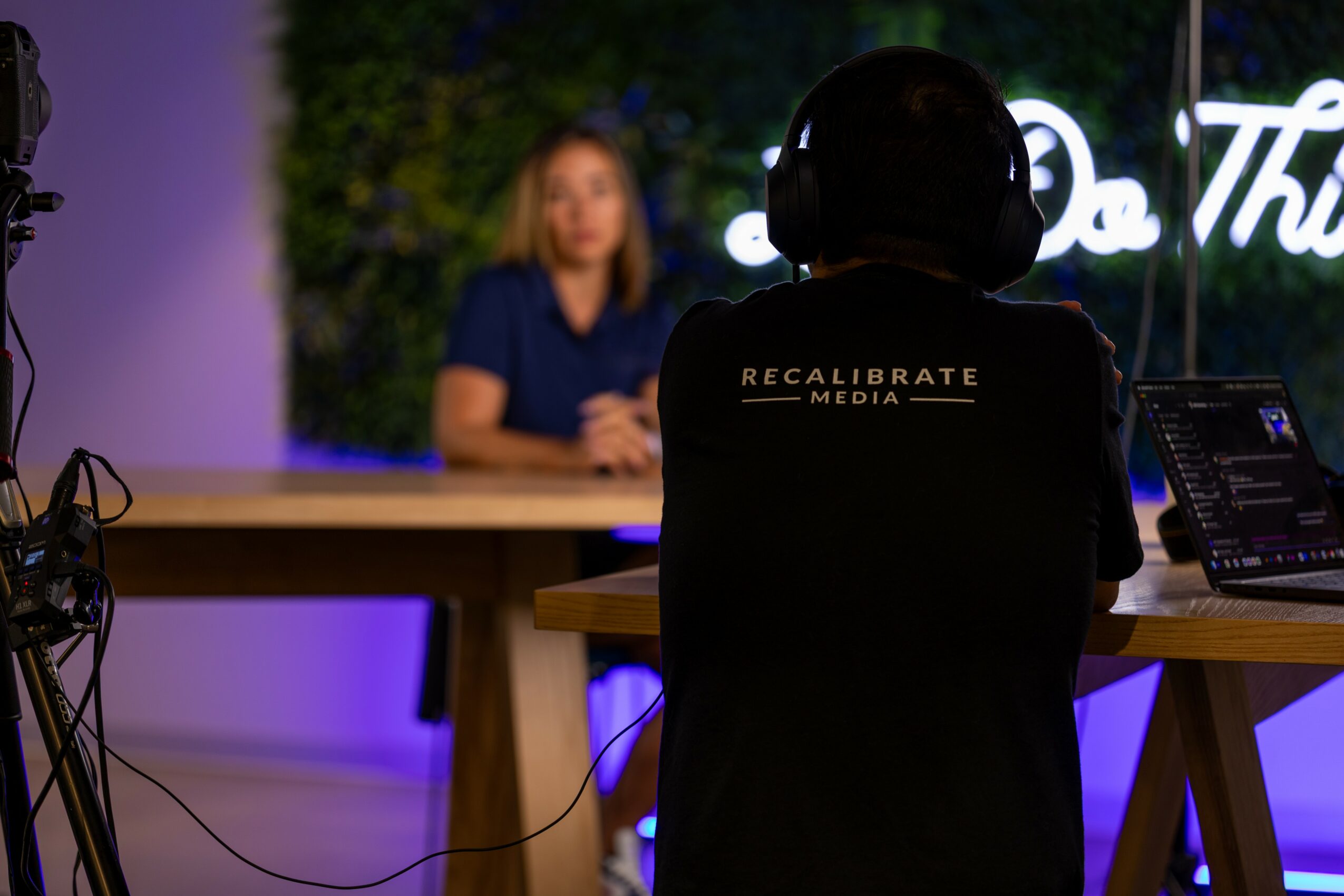 Person wearing a helmet and a jacket with 'Recalibrate Media' sits at a desk with a laptop, facing a woman at a table.