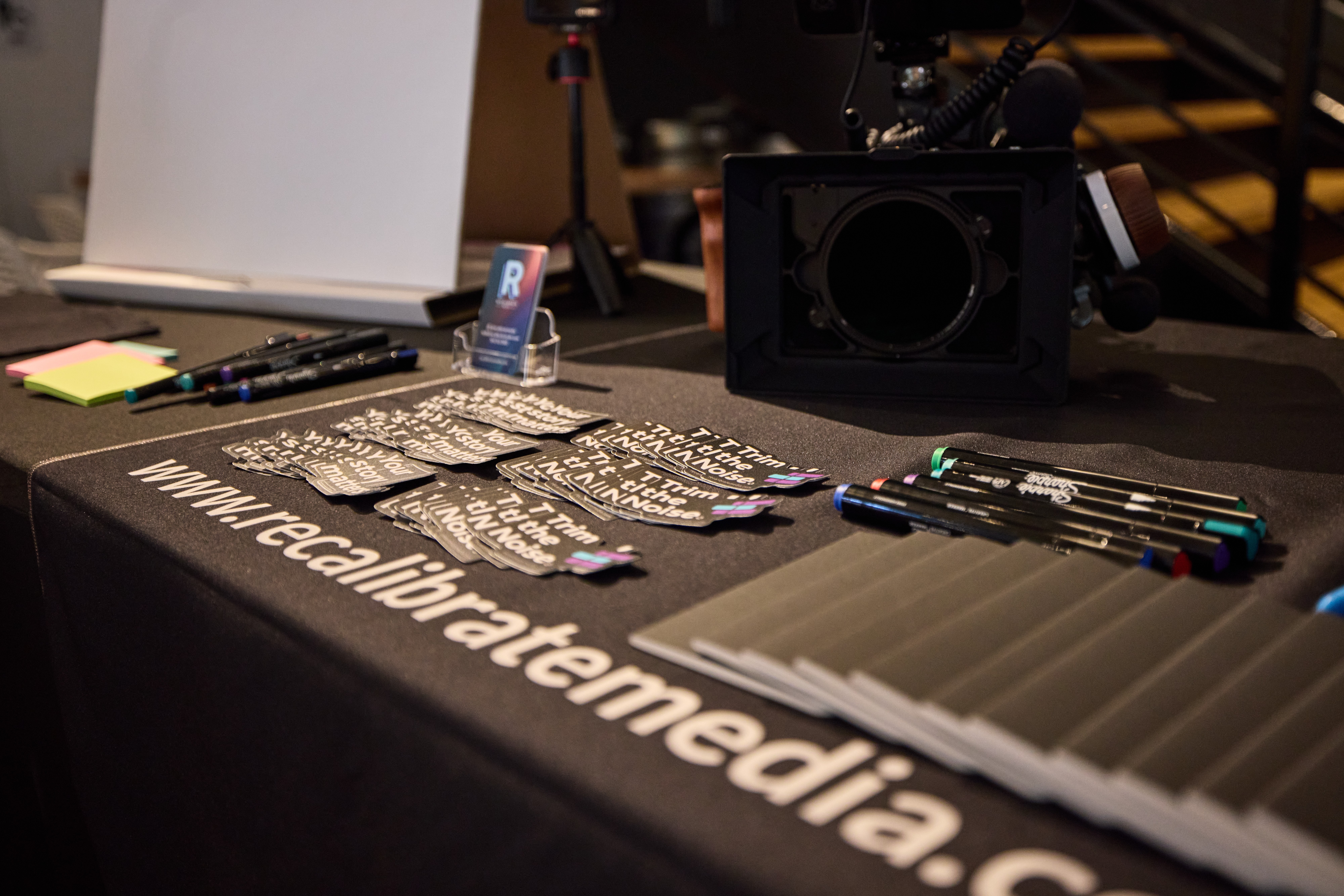 Table with promotional items, pens, and a camera, with a website URL printed on the tablecloth.