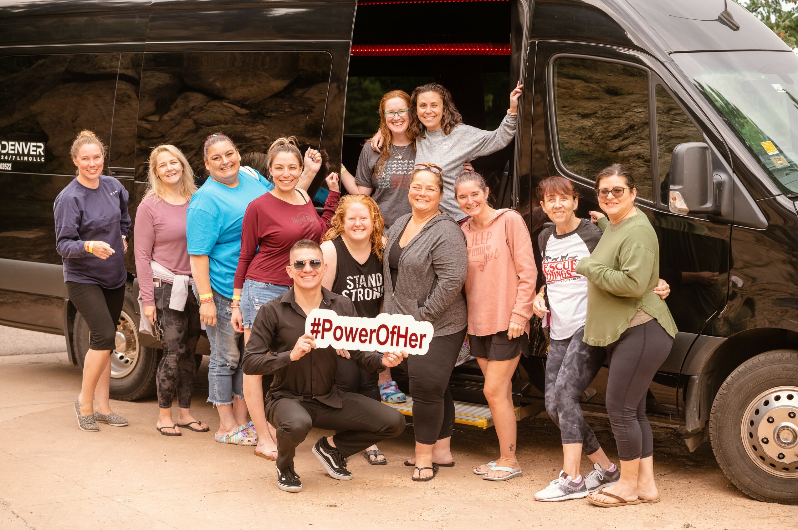 Group of people standing in front of a black van, holding a #PowerOfHer sign, smiling outdoors.