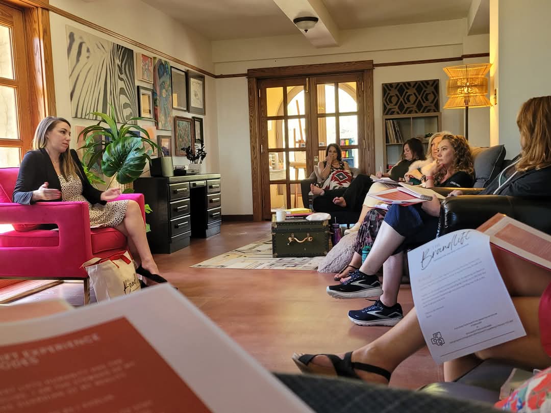 Group of women sitting in a room during a discussion or meeting, with artwork on the wall and large windows in the background.