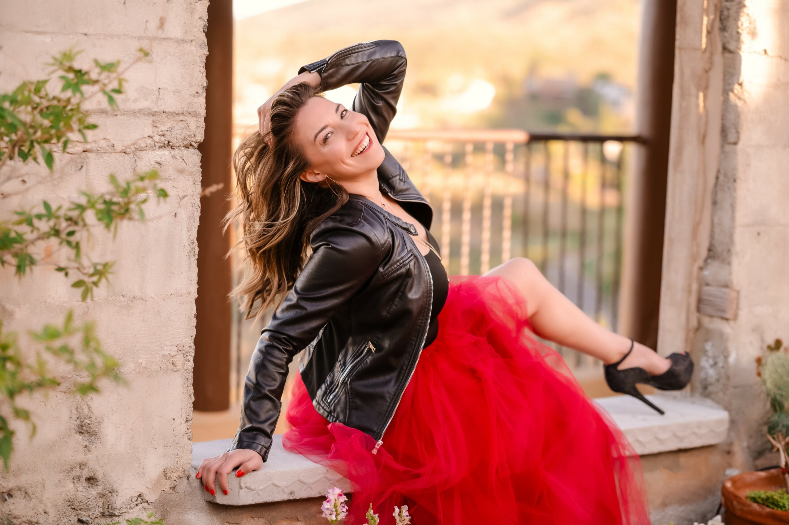 Woman in black leather jacket and red skirt sitting on a stone ledge, smiling, with a blurred outdoor background.