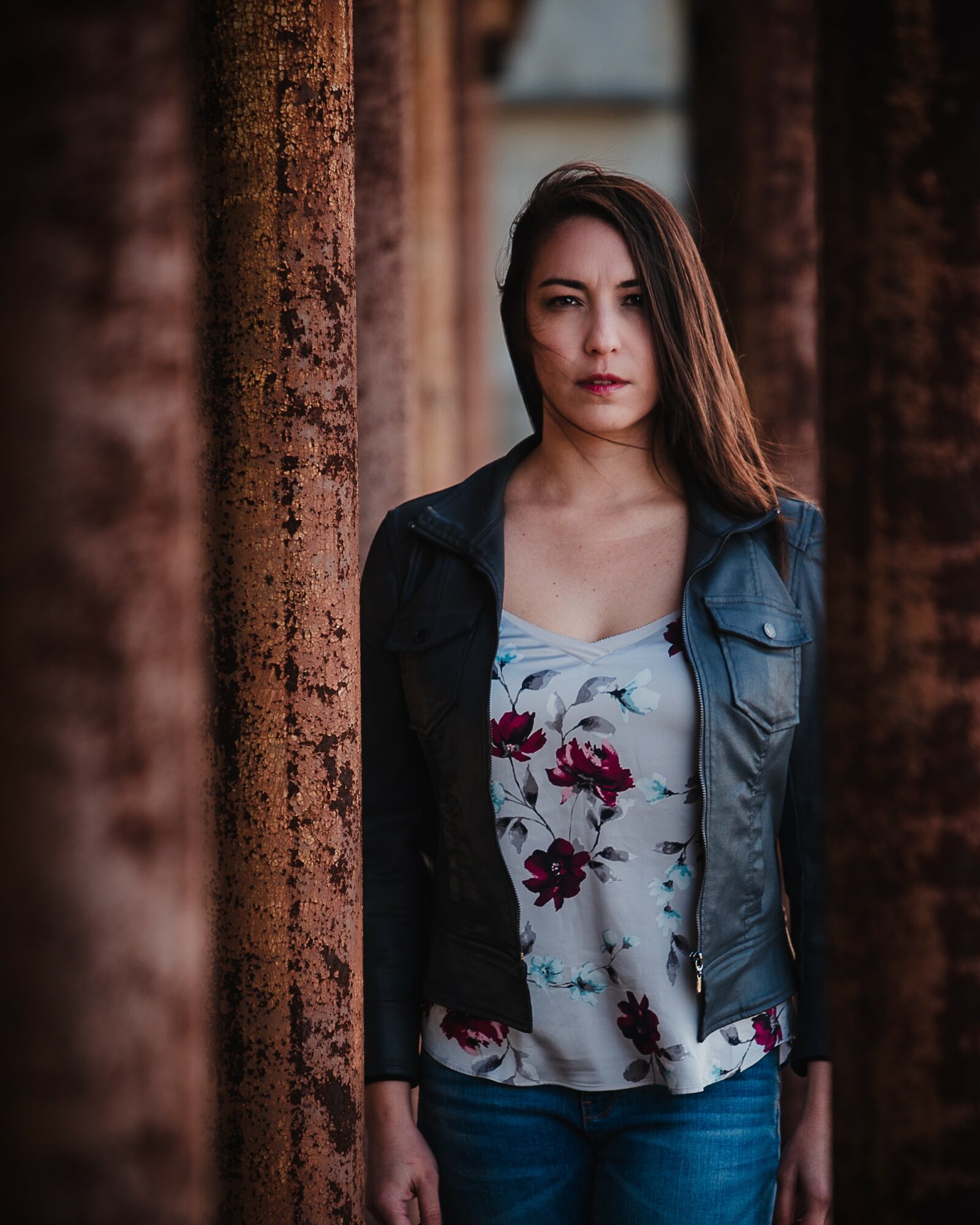 Woman with long hair standing between rusted metal beams outdoors, wearing a floral top and leather jacket.