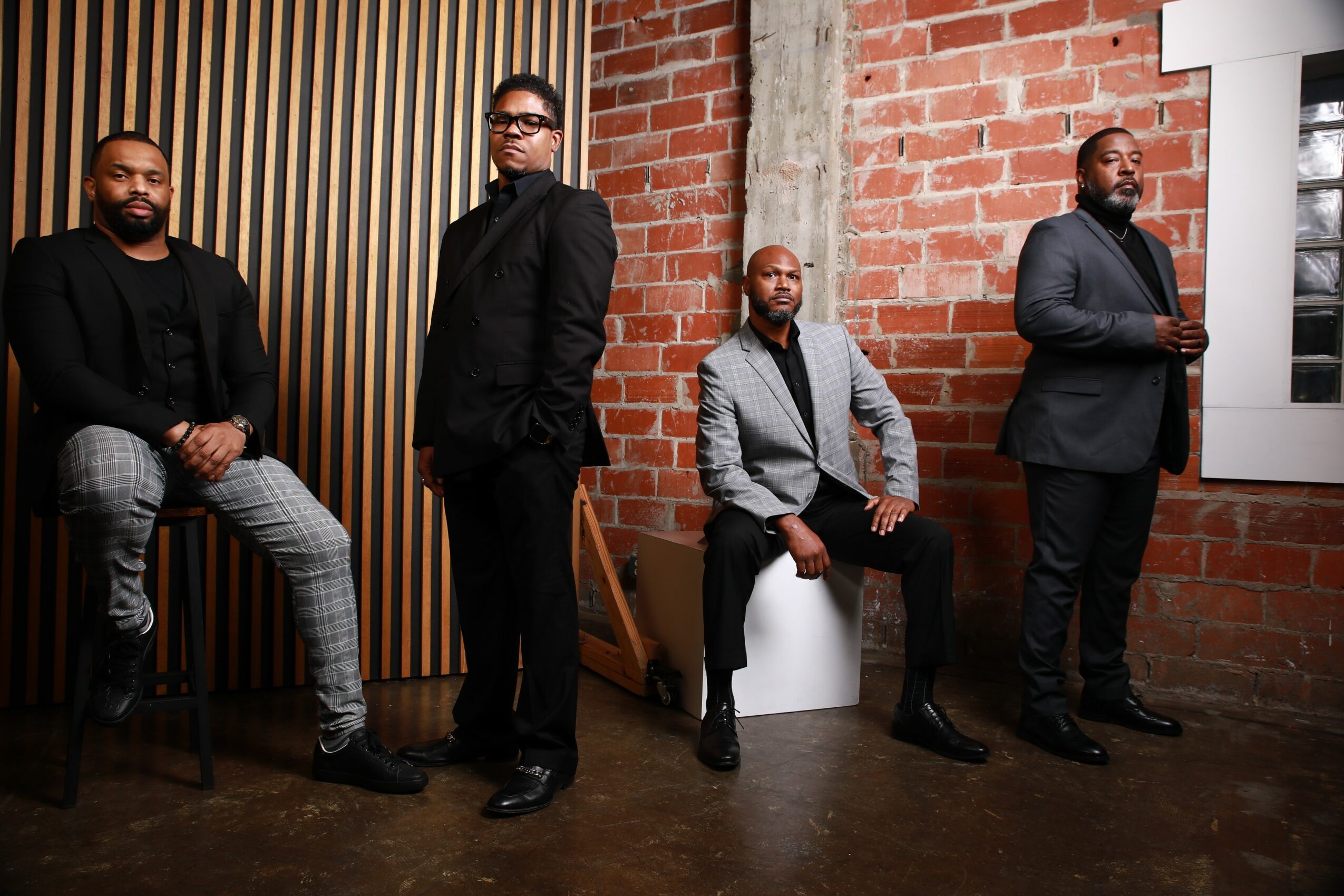 Four men in formal attire pose against a brick wall and wooden panel background, some sitting and some standing.
