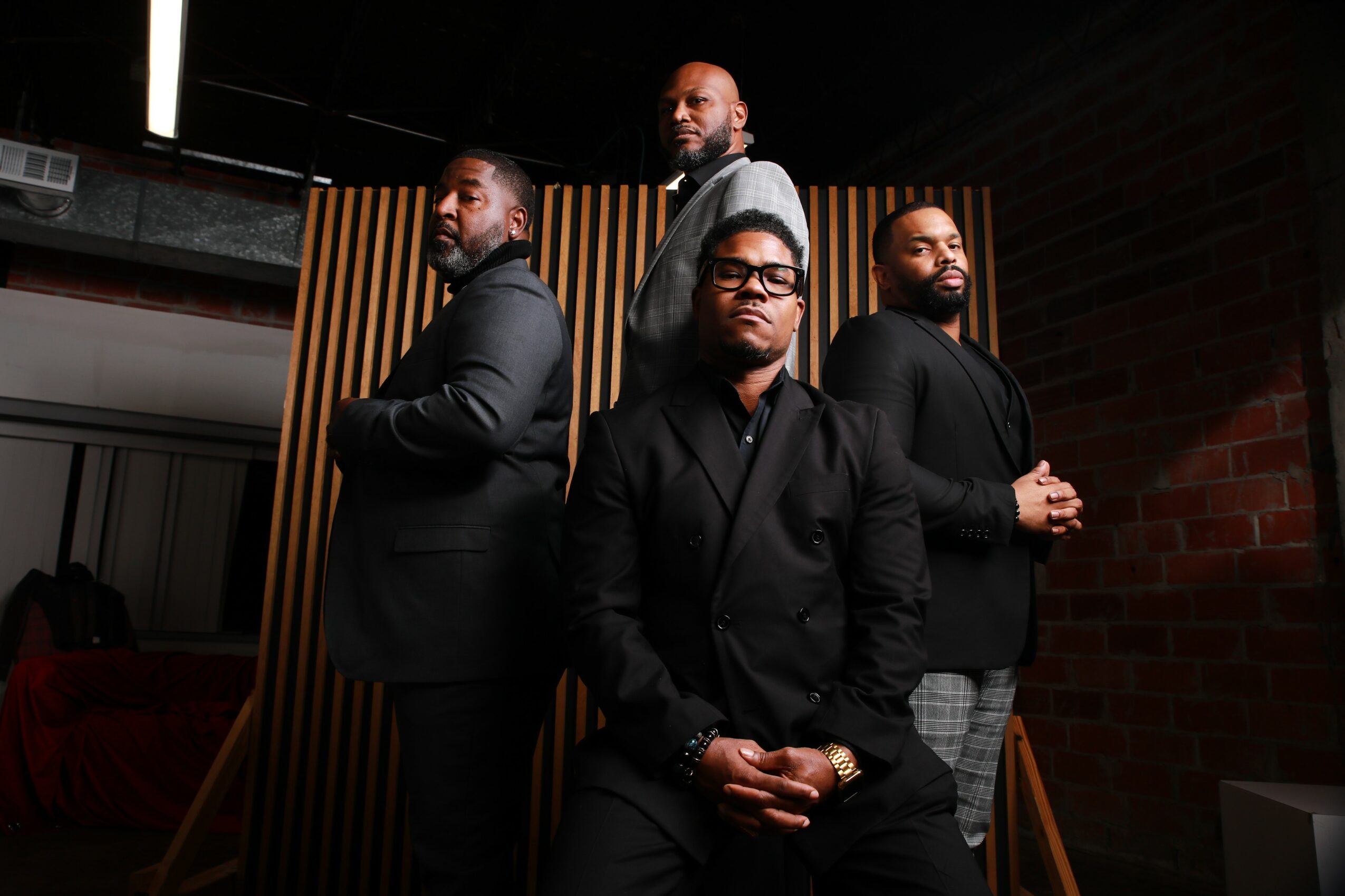 Four men standing in a row against a wooden and brick background, dressed in dark clothing, looking serious.