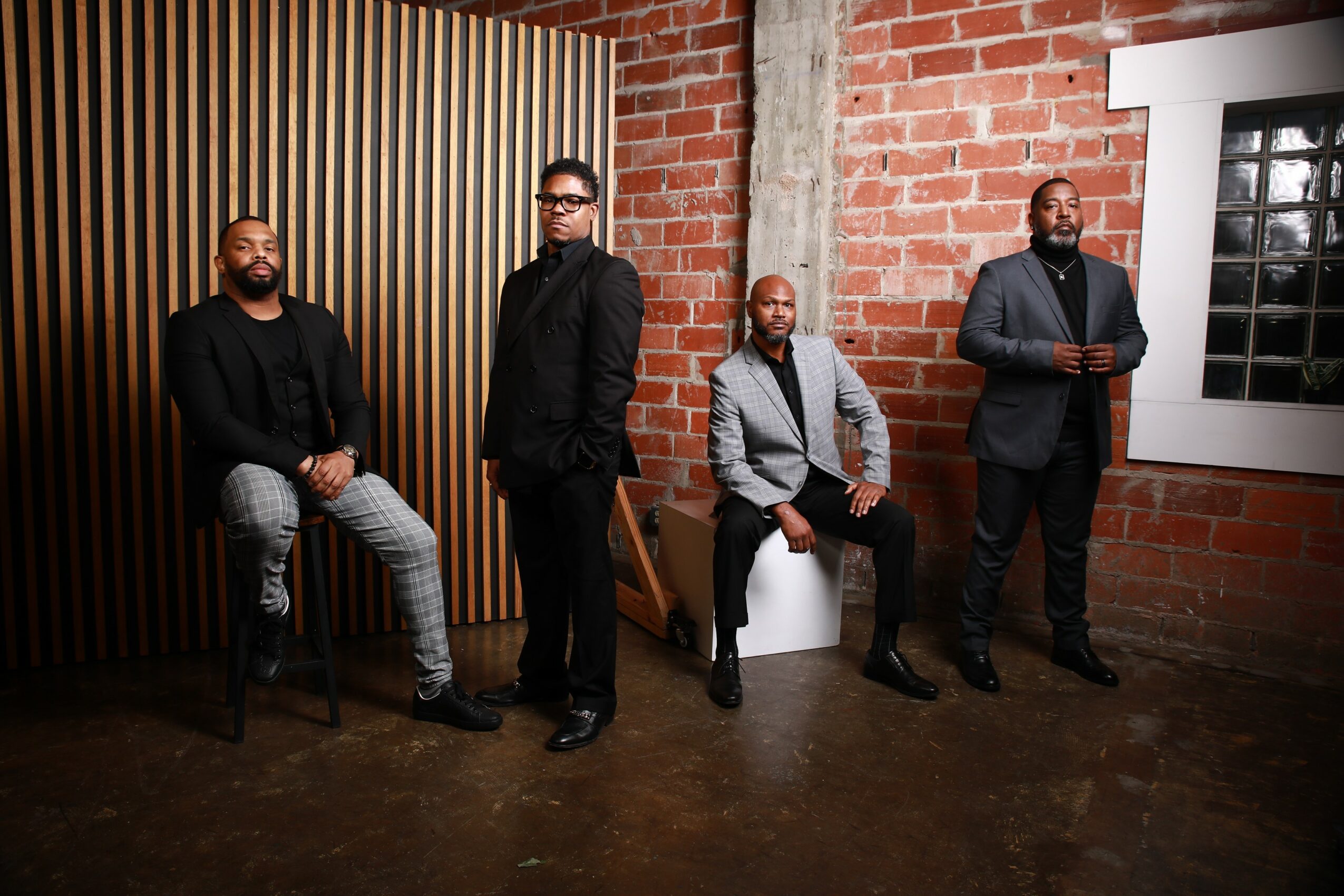 Four men in formal and casual attire pose in an industrial-style room with brick walls and wooden paneling.