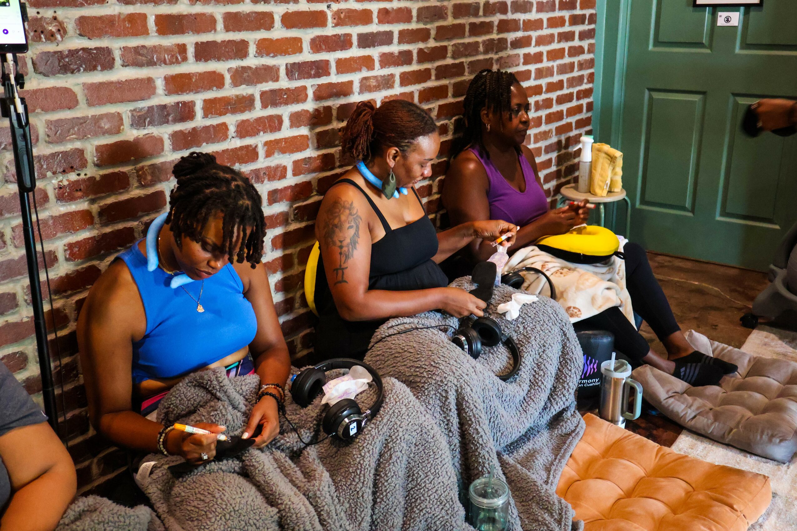Three women sit on a bench against a brick wall, knitting and chatting, with bags and drinks nearby.