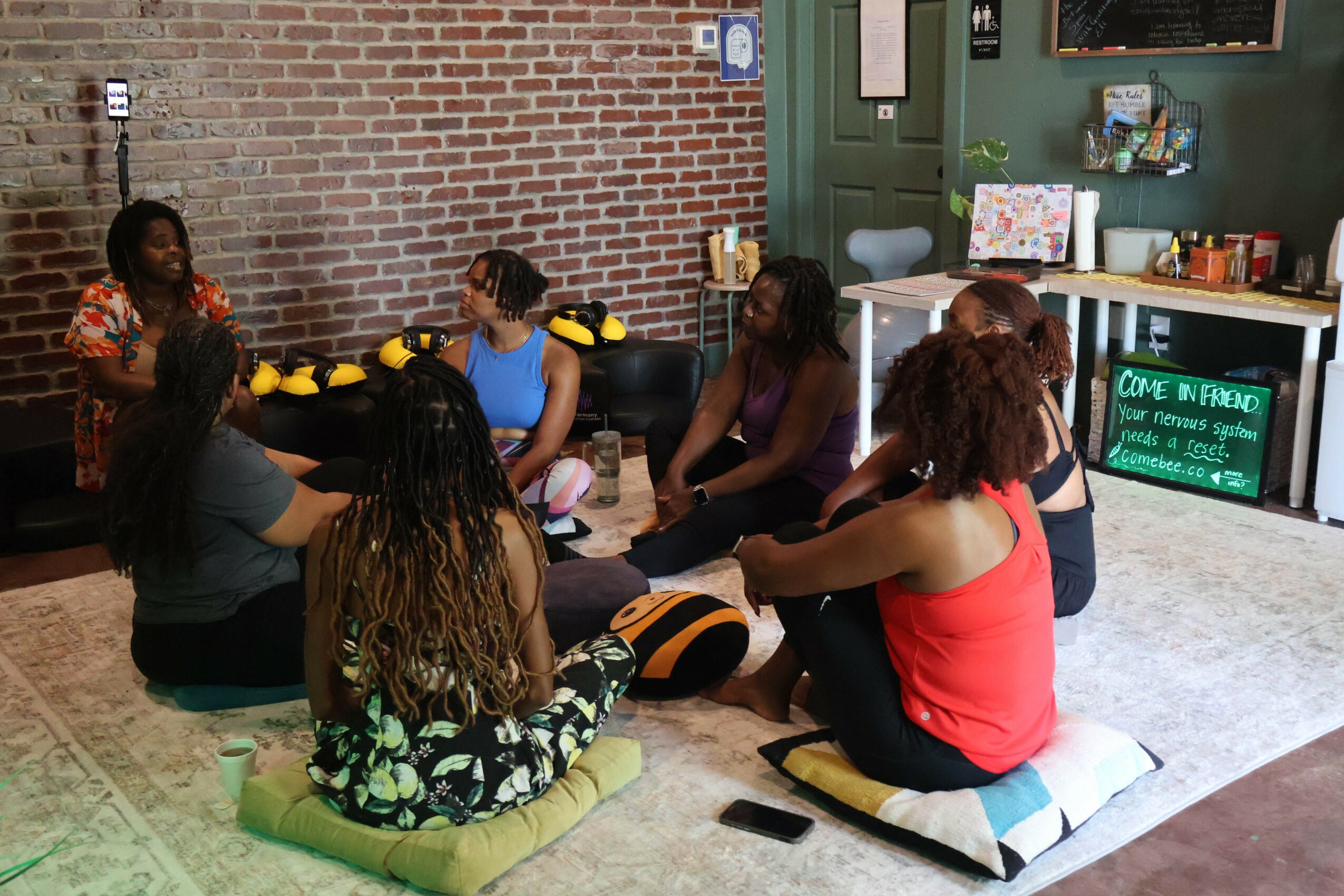 Group of people sitting on cushions and chairs in a room with brick wall, listening to a woman speaking, with books and a chalkboard in the background.