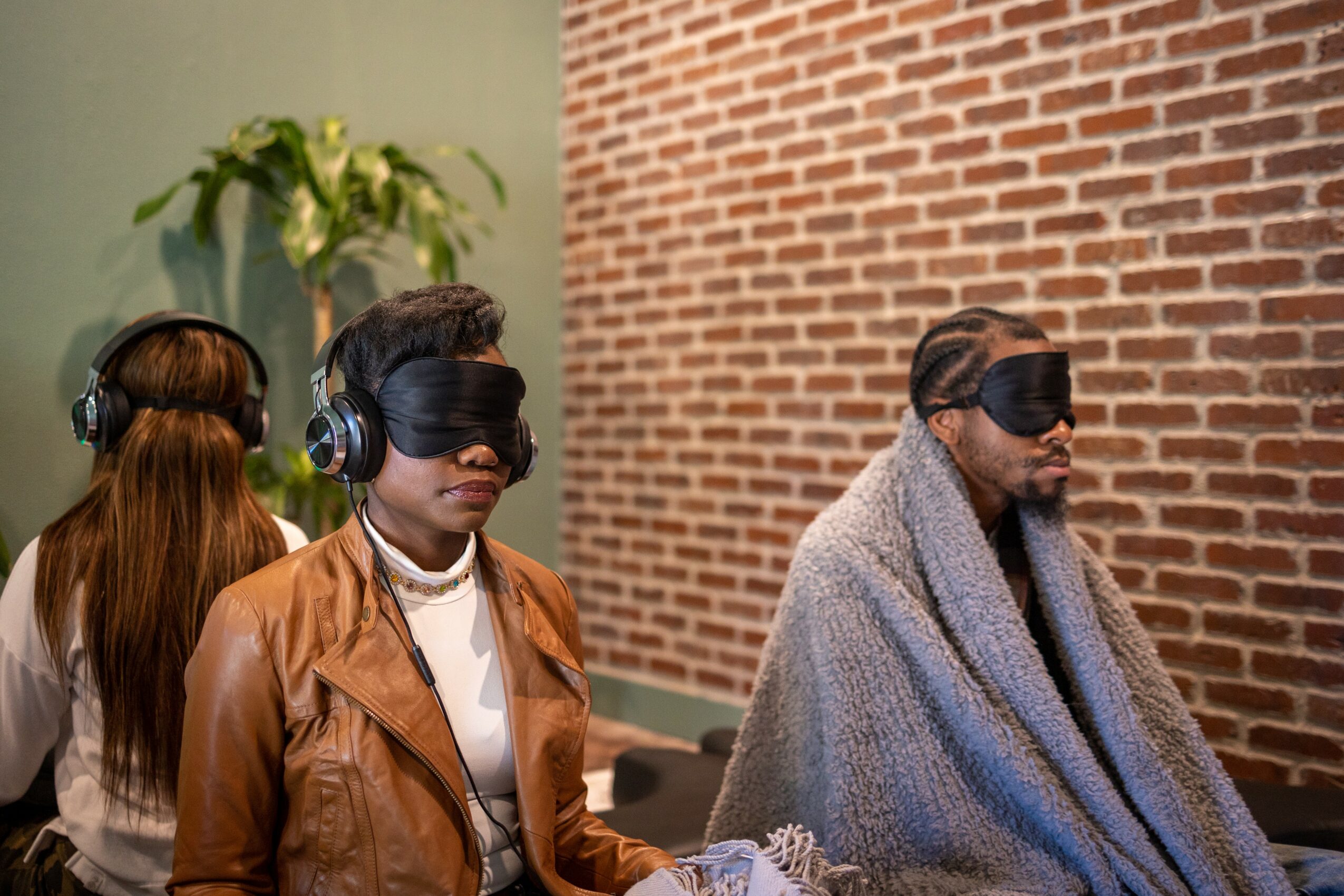 Three people sit against a brick wall, wearing blindfolds, with one person wrapped in a blanket, in an indoor setting.