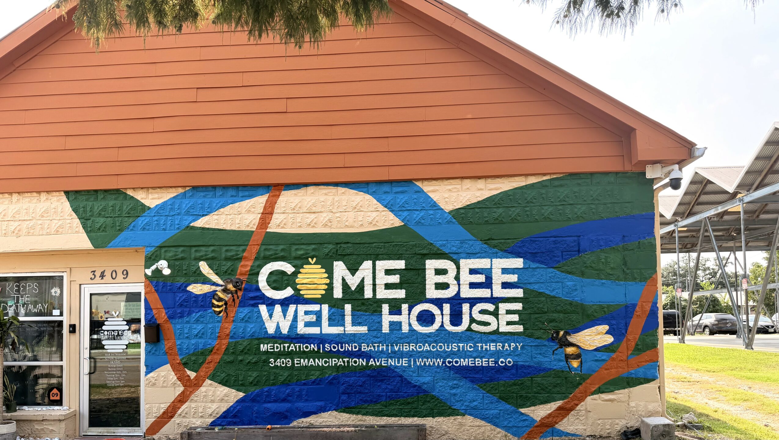 Colorful mural on building wall with text 'Come Bee Well House' and bee graphic, featuring blue, green, and orange waves.