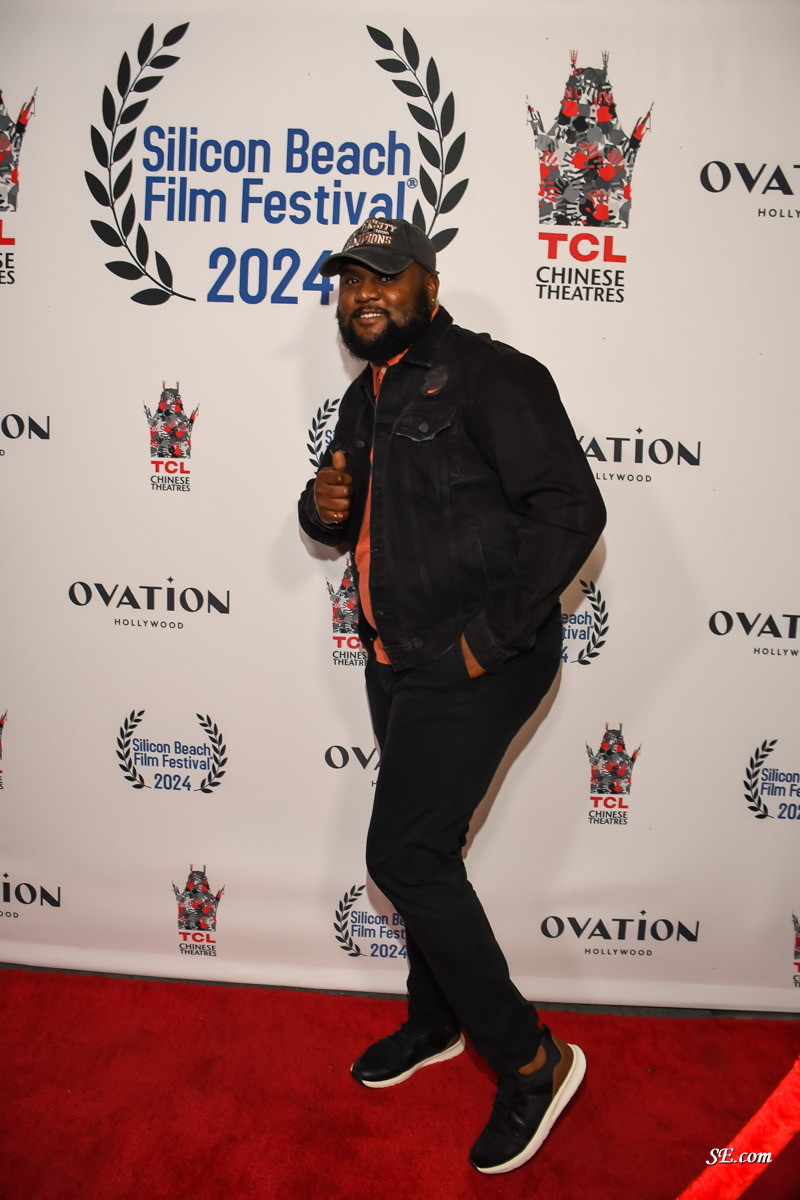 Man in black jacket and cap standing on red carpet at Silicon Beach Film Festival 2024, smiling, giving thumbs up.