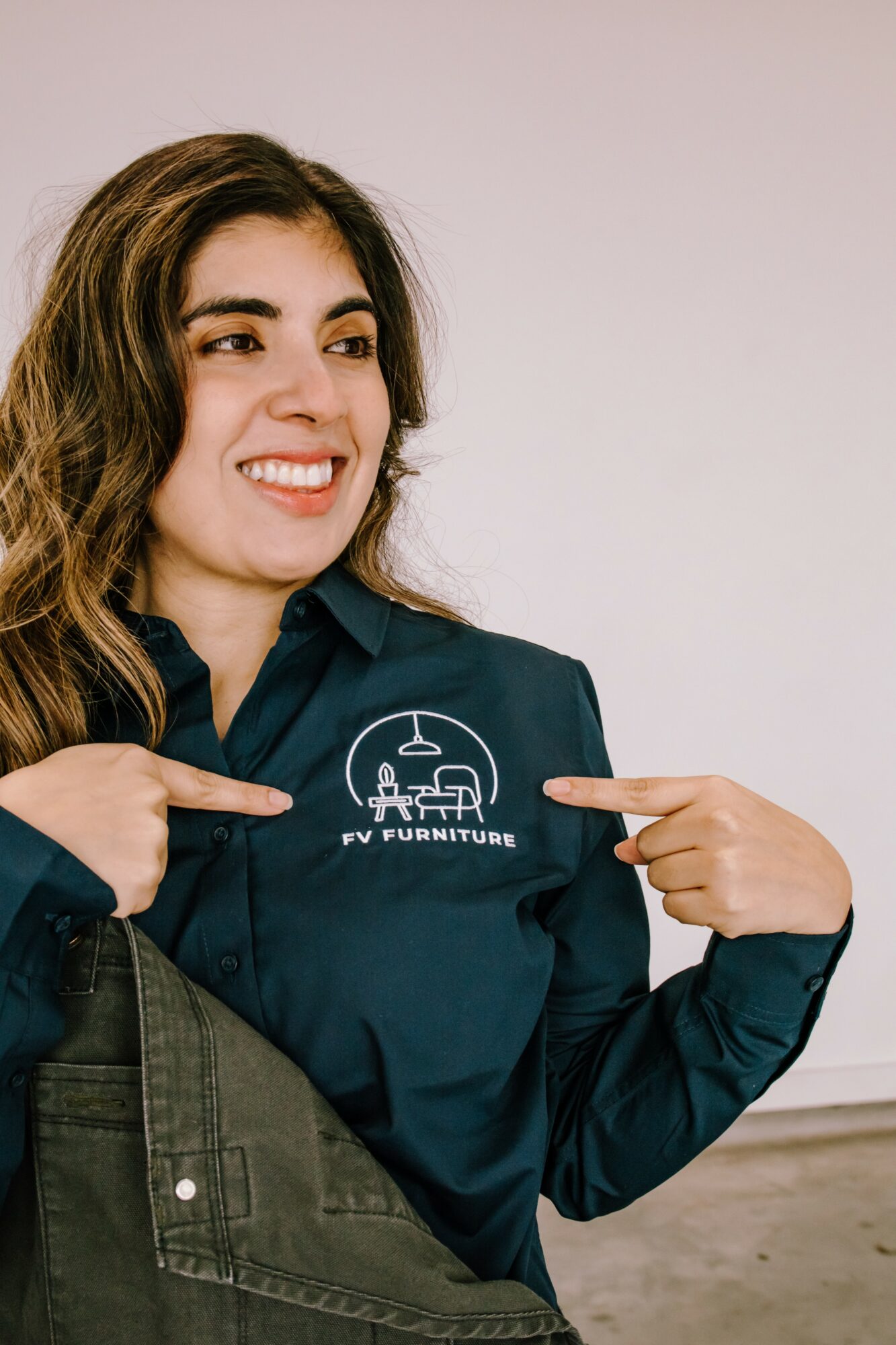 Woman smiling and pointing at logo on her shirt, holding a bag, against plain background.