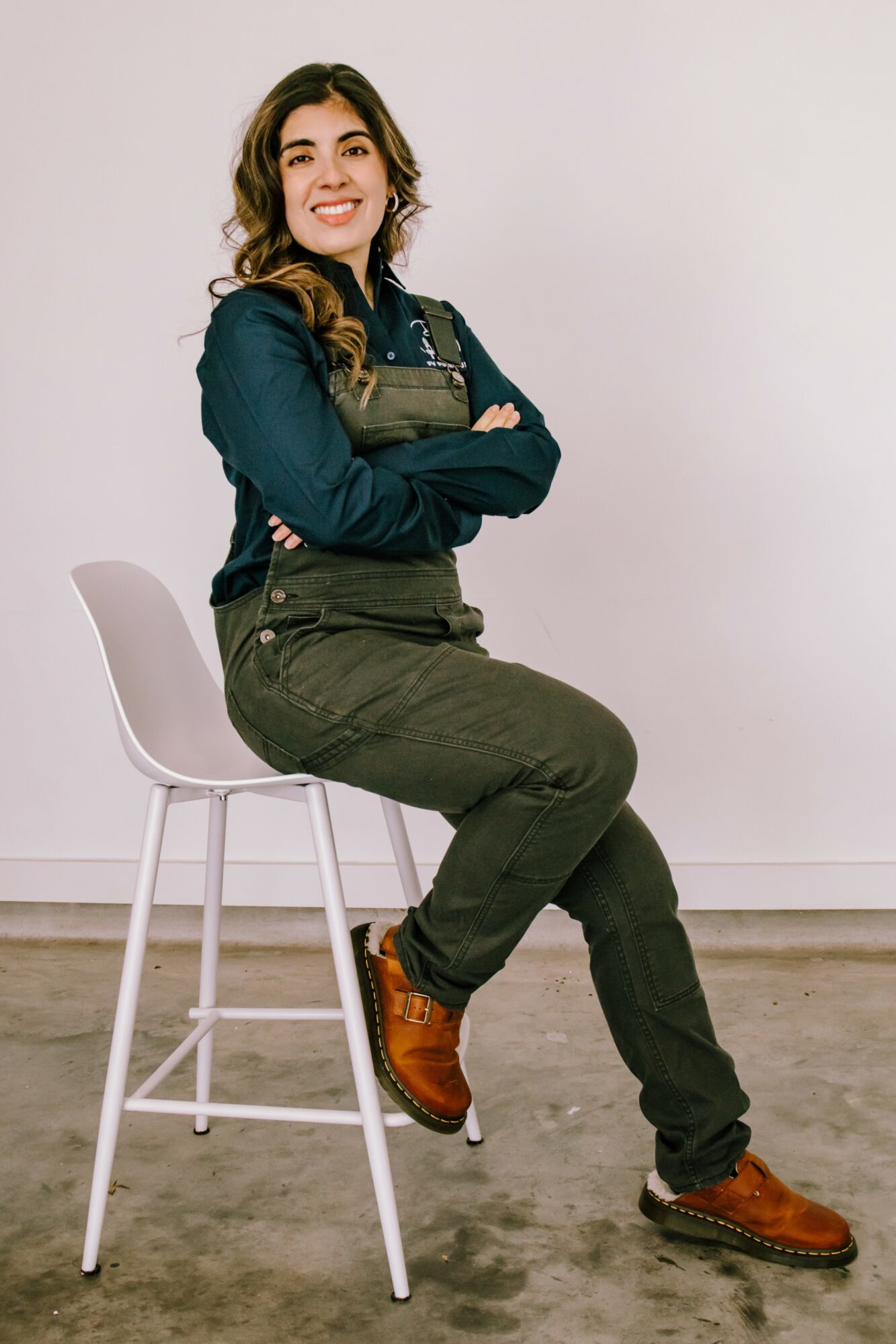 Young woman sitting on a white chair, smiling, arms crossed, against a plain white background.