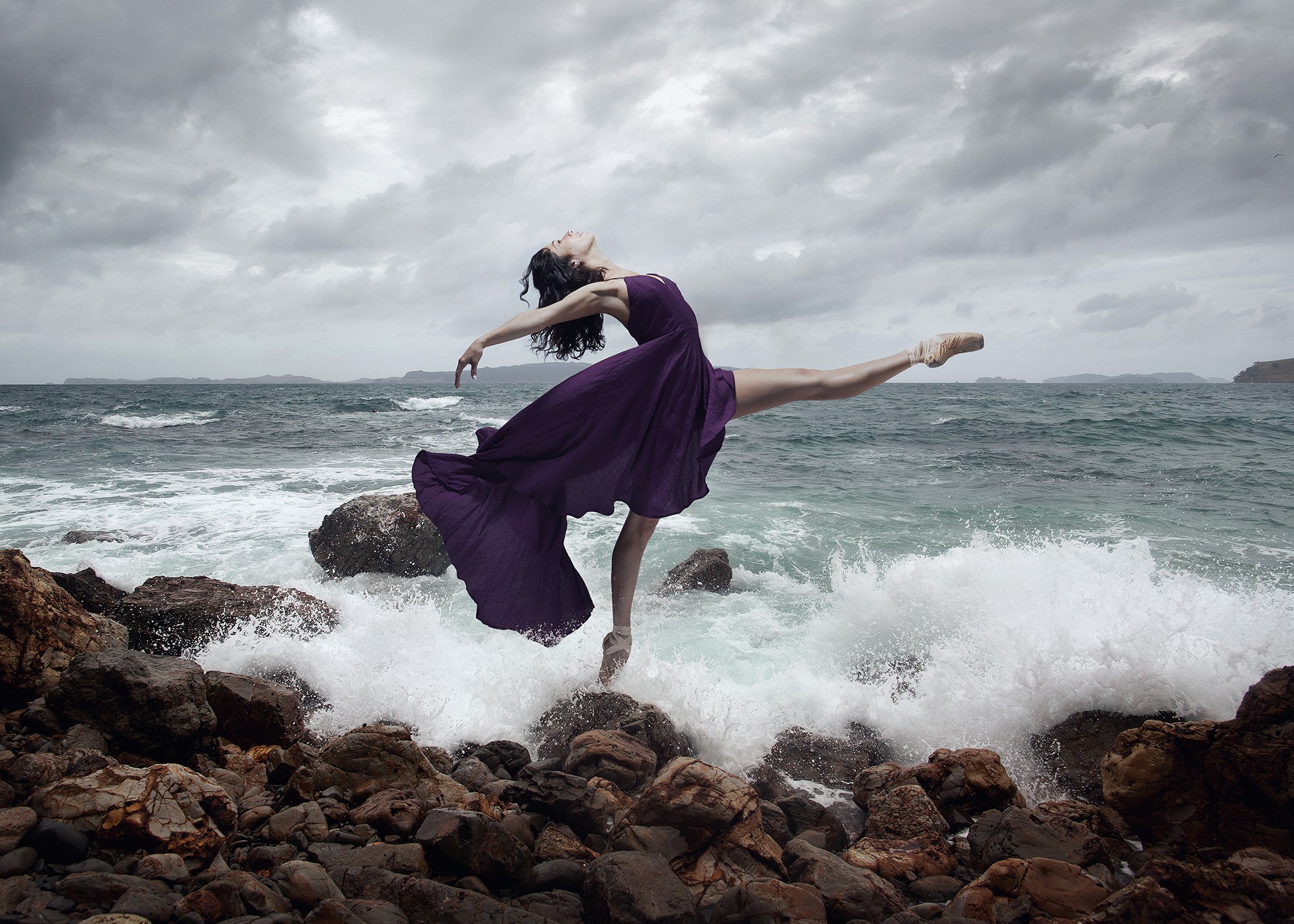 Person in a purple dress balancing on rocks by the ocean, with waves crashing around them and cloudy sky above.