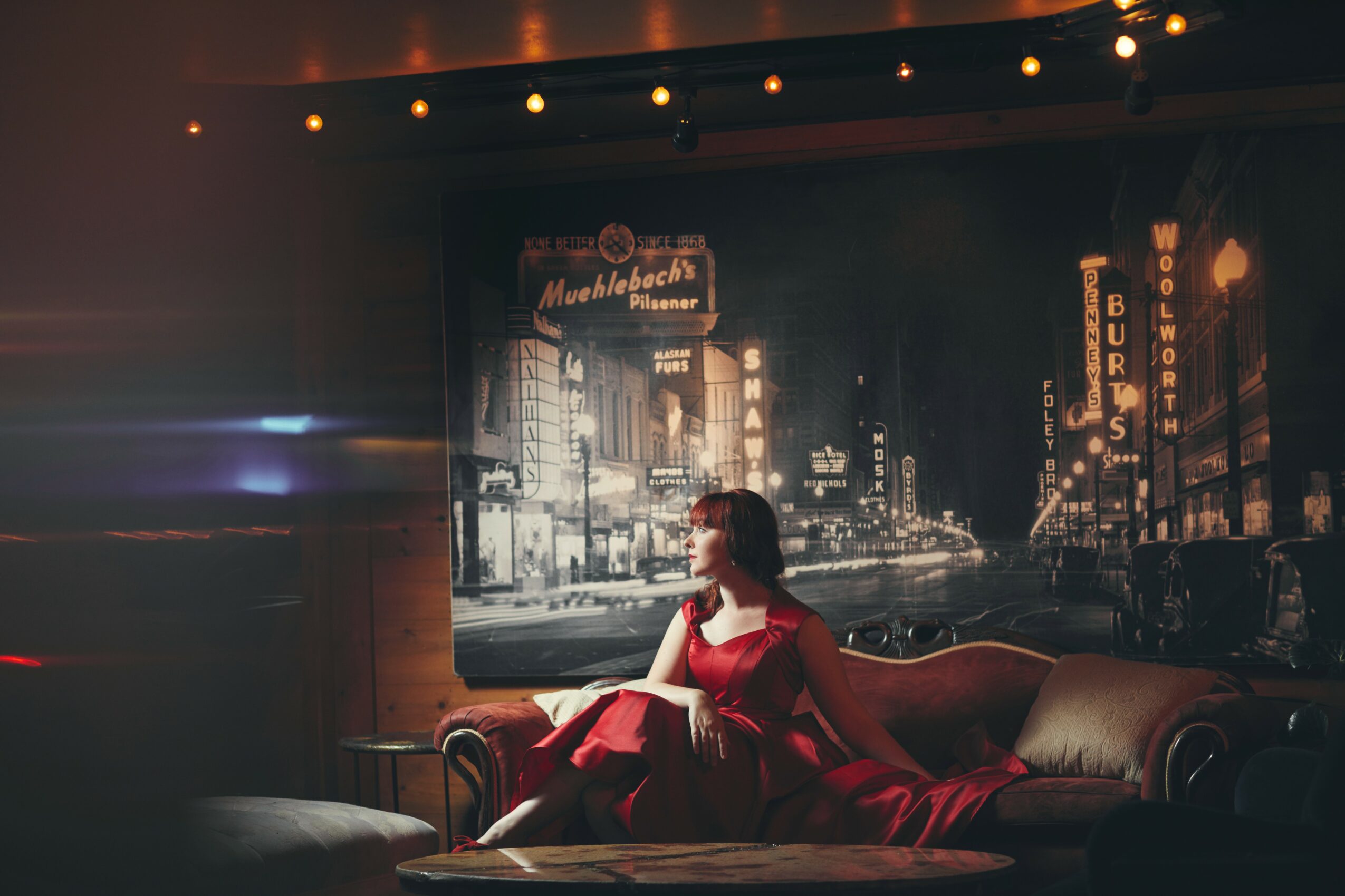 Woman in red dress sitting on a vintage sofa in a dimly lit room with cityscape mural background.