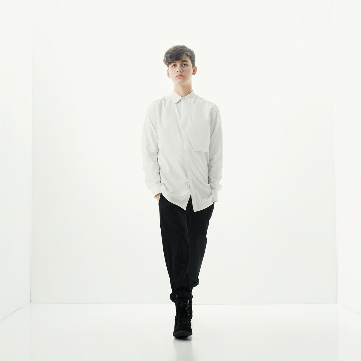 Young man walking in a white room, wearing a white shirt and black pants, with hands in pockets.