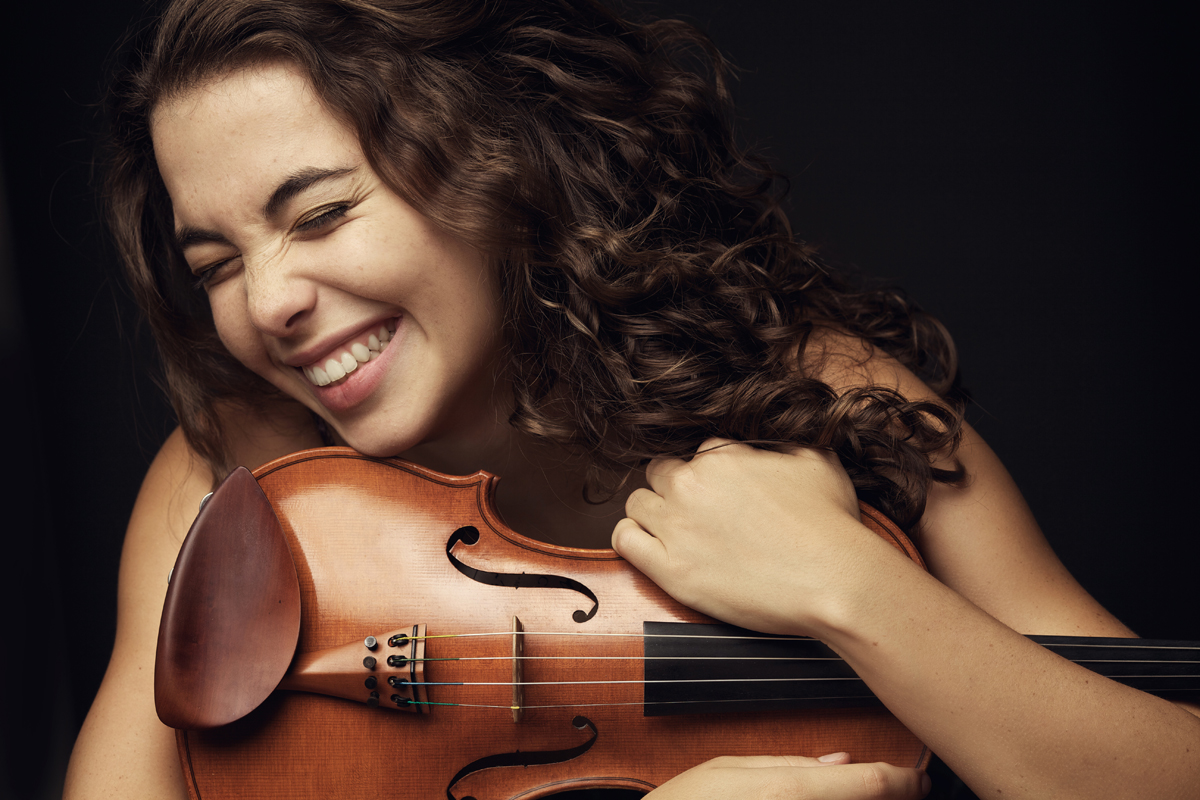 Woman with curly hair smiling and holding a violin, dark background, close-up.