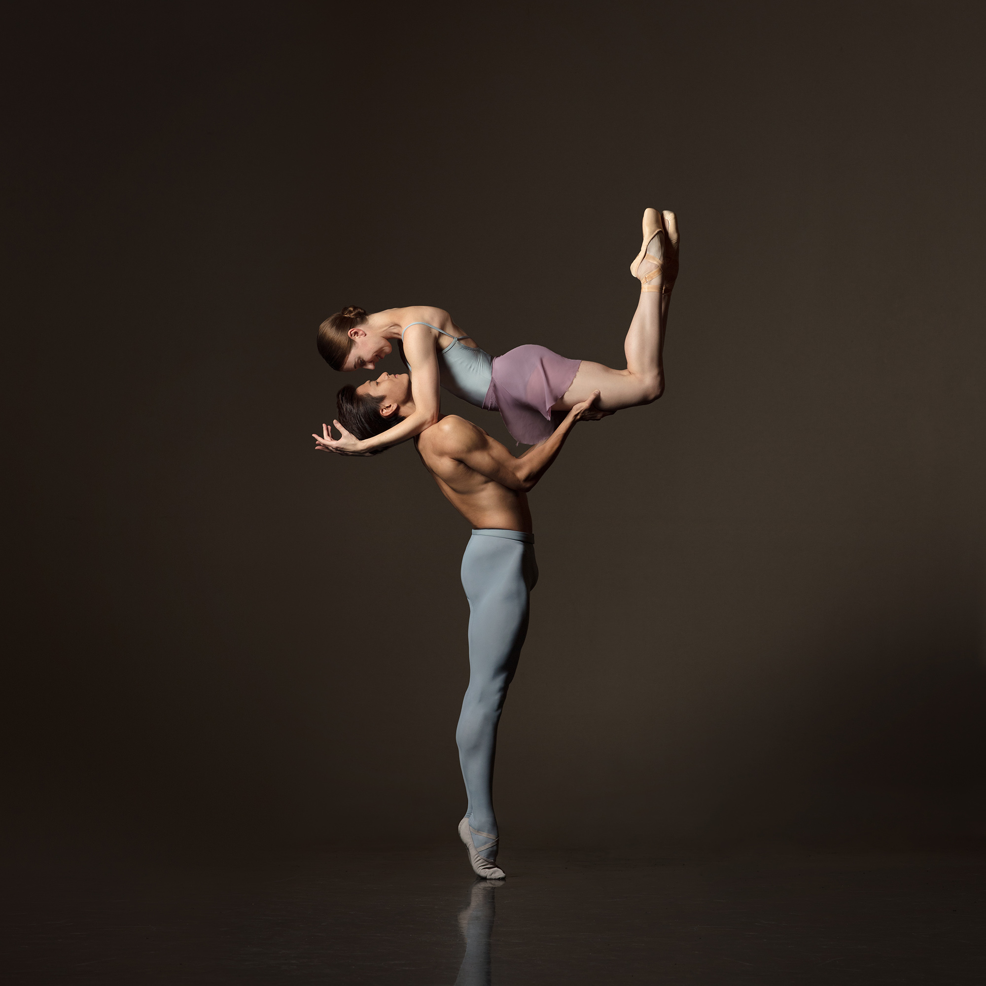 Two ballet dancers perform an acrobatic pose against a dark background, with one supporting the other in a lift.