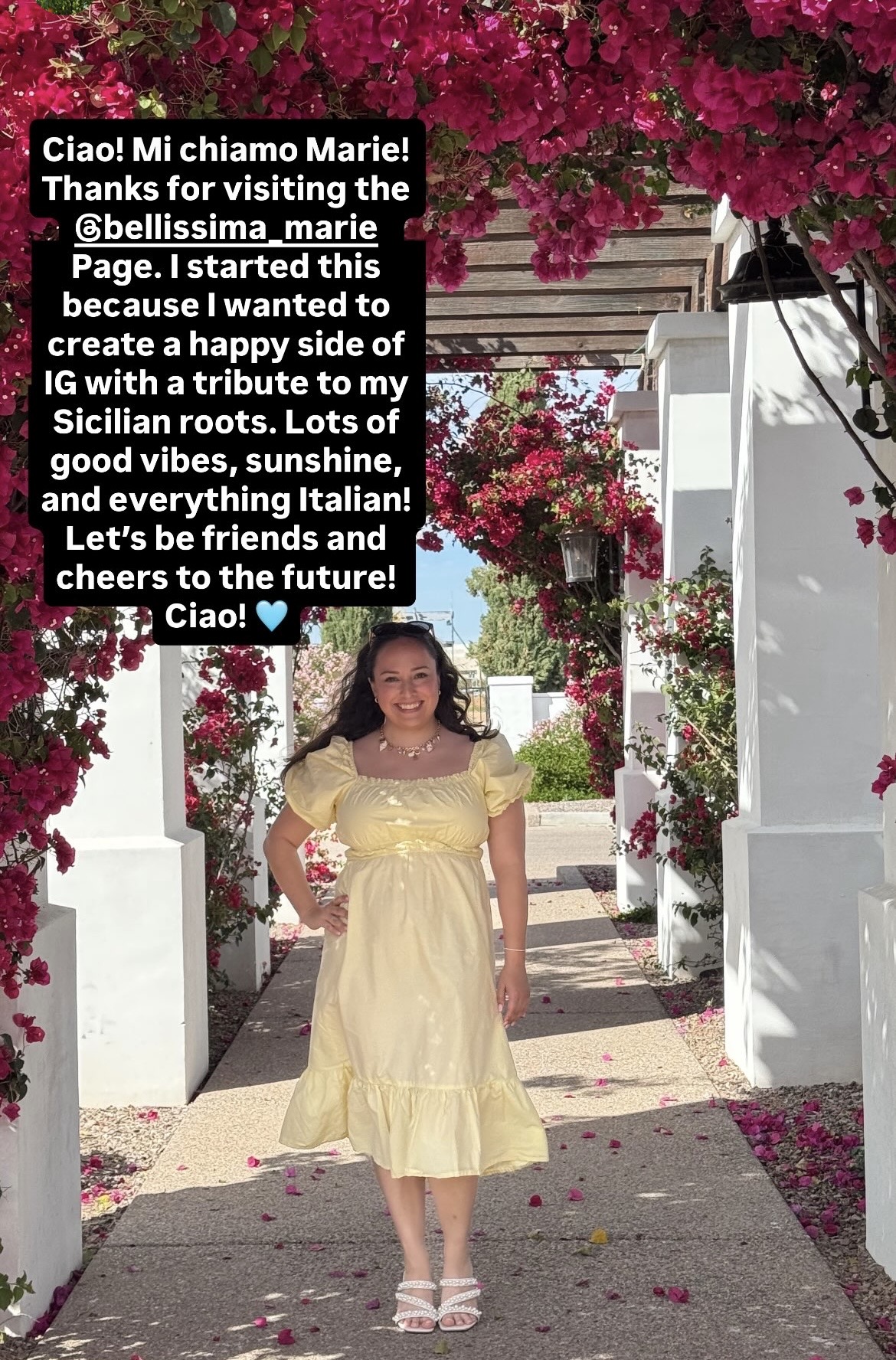 Young girl in a yellow dress standing on a pathway with white arches and pink flowers, holding a bouquet, smiling.