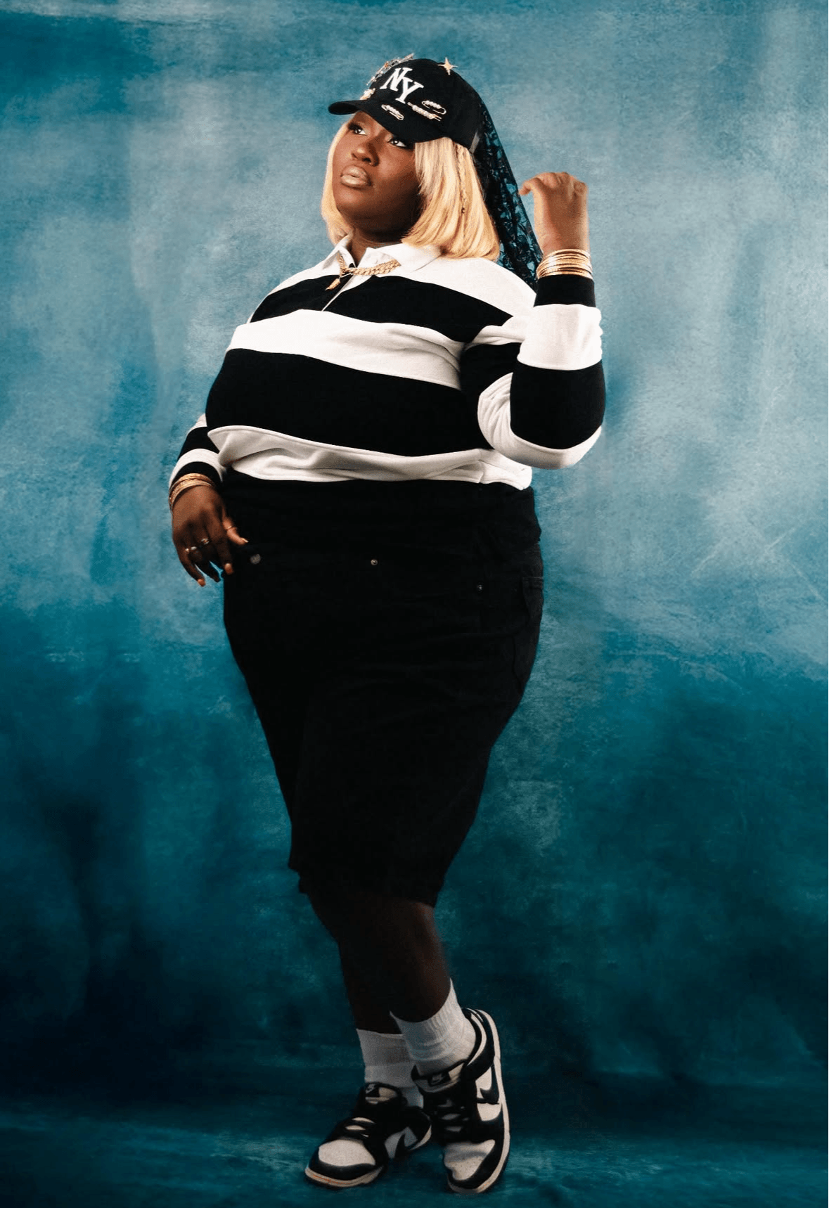 Person with blonde hair wearing a black cap, striped black and white shirt, black skirt, and sneakers, standing against a blue background.