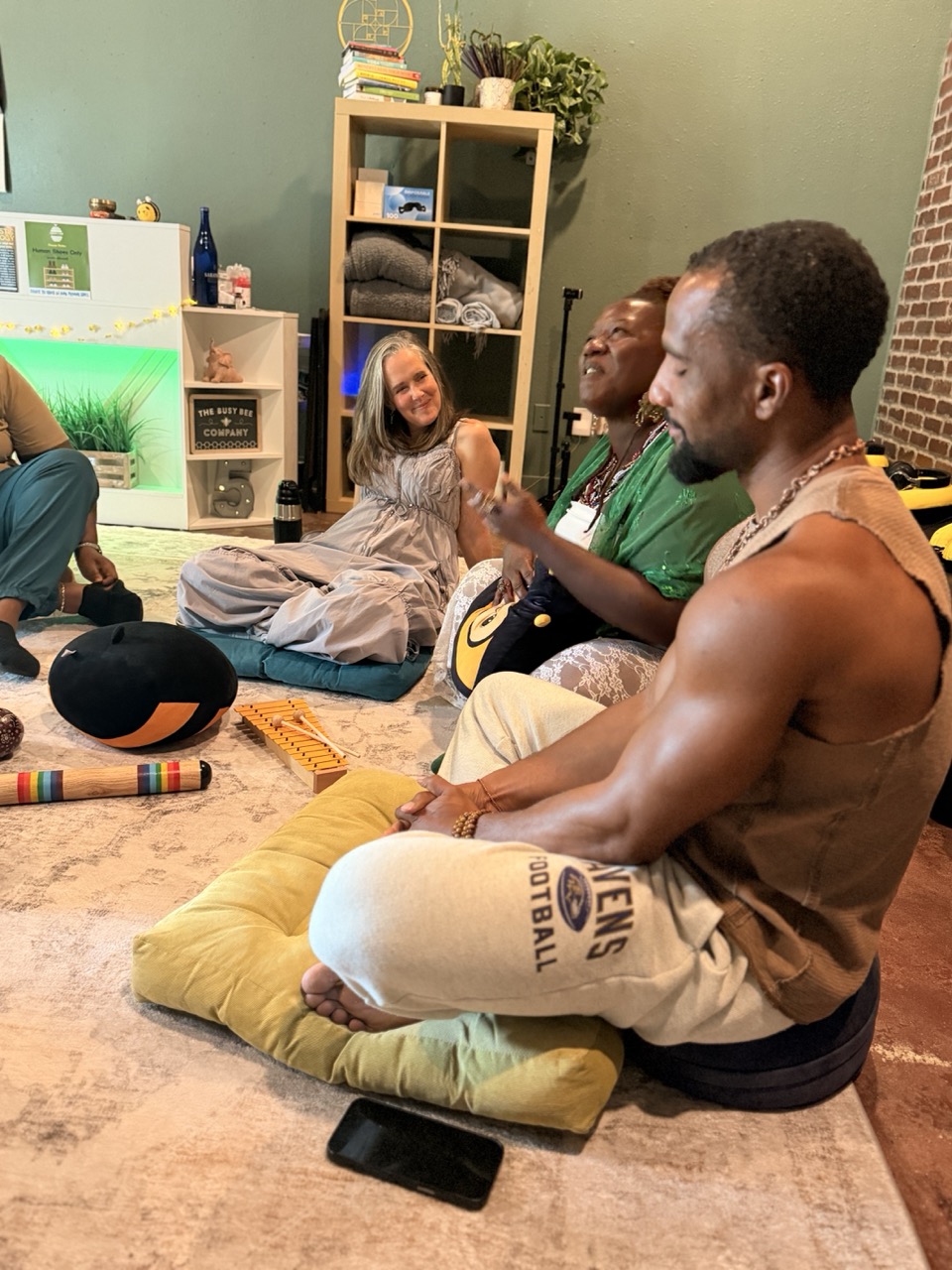 Four people sitting on cushions and a blanket on the floor, engaging in conversation in a cozy room with shelves and plants.