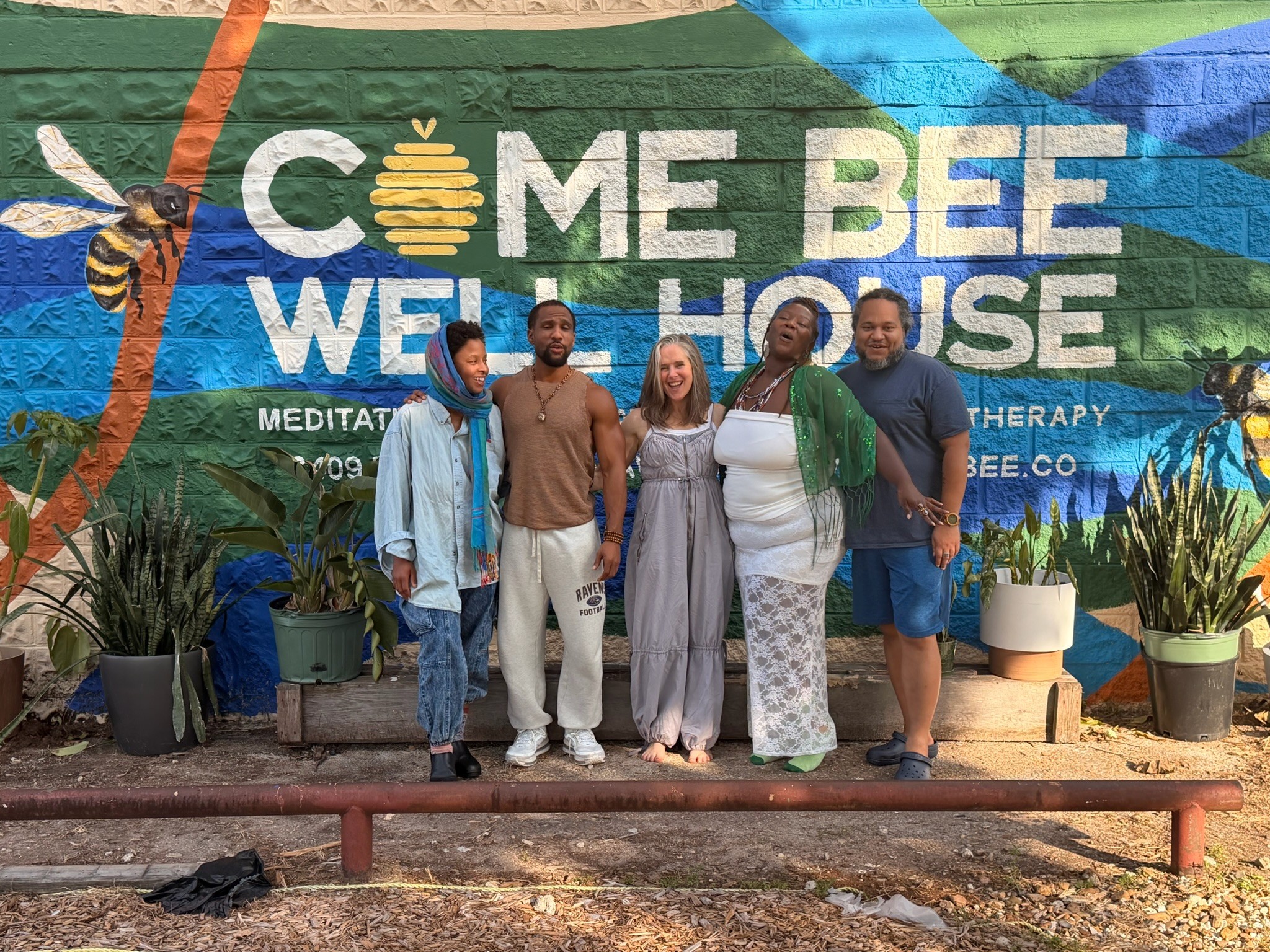 Group of five people standing in front of a colorful mural with text about a bee-themed wellness house, plants in pots nearby.