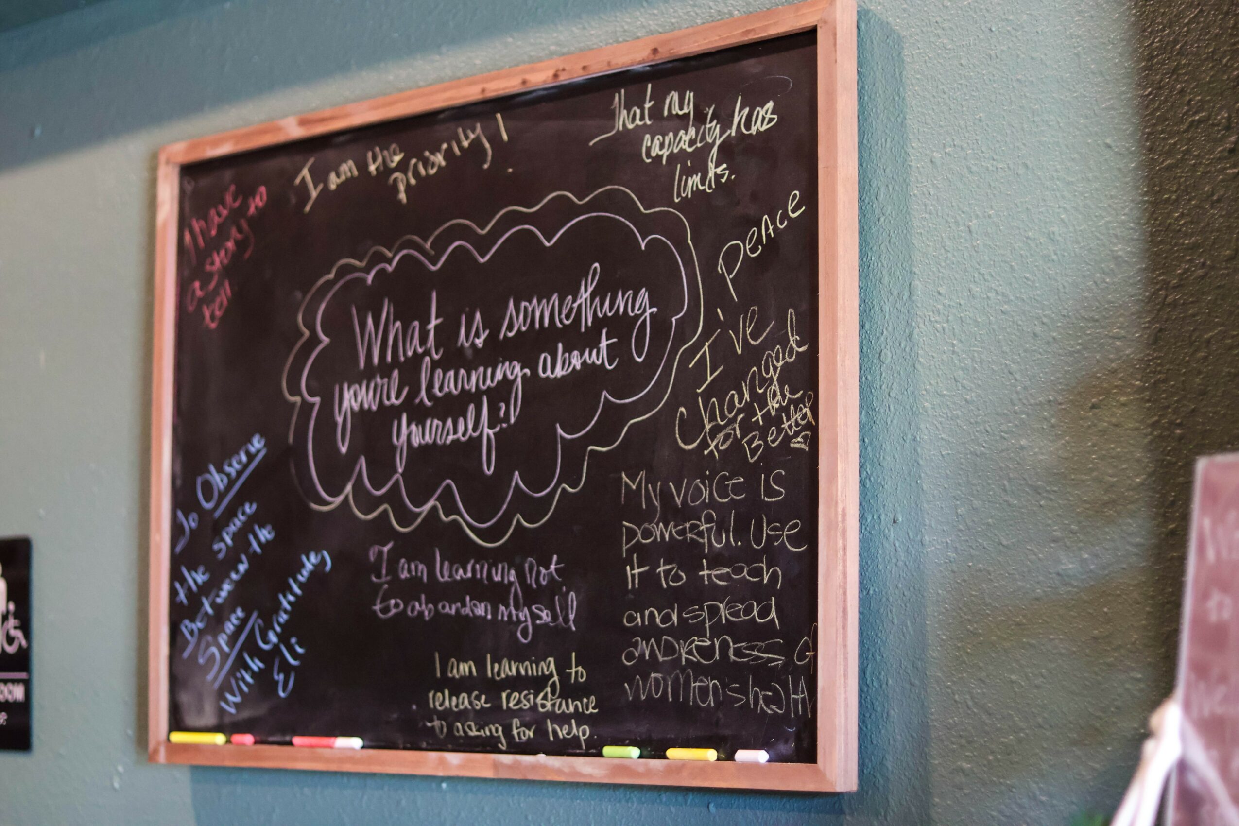 Chalkboard with handwritten quotes and messages, framed in wood, on a wall. Partially visible pink object on the right.