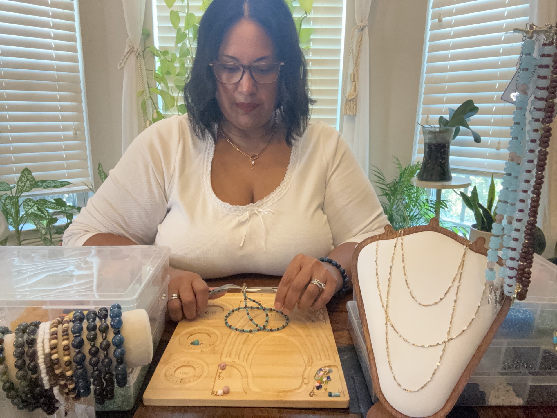 Woman with glasses sitting at a table with jewelry display, plants, and window blinds in background.