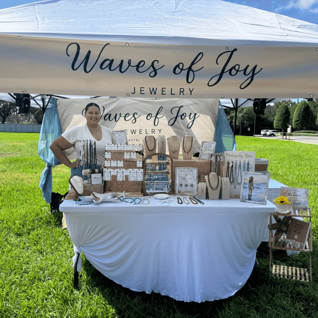 Woman standing behind a jewelry booth under a tent with a sign reading 'Waves of Joy Jewelry' in a park.