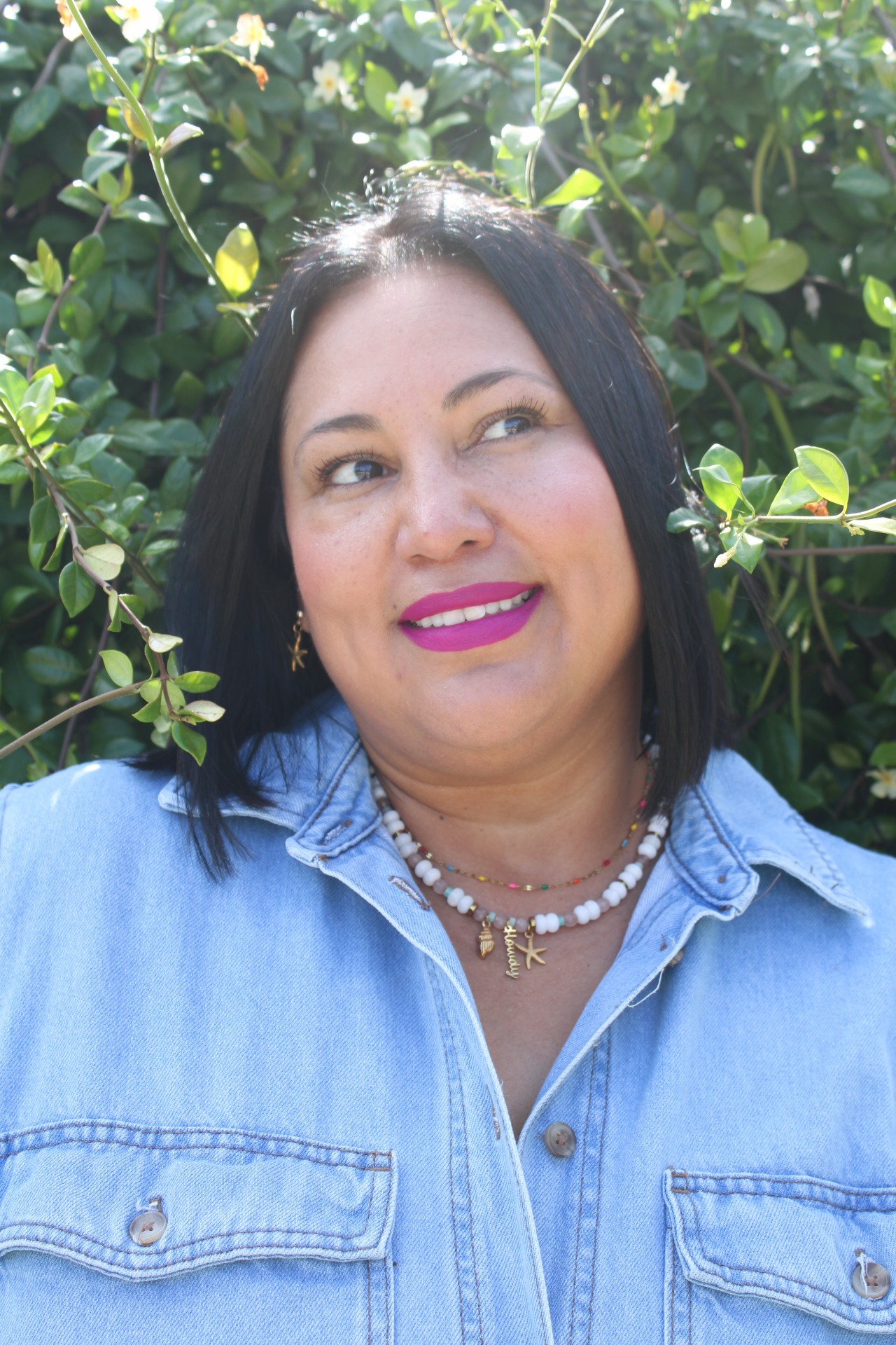 Woman with black hair and pink lipstick smiling outdoors, wearing a denim shirt and layered necklaces, surrounded by green foliage.