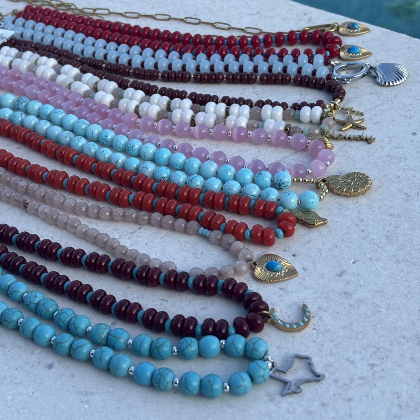 Multiple colorful beaded necklaces with various charms, arranged on a light surface, featuring red, blue, pink, white, and purple beads.
