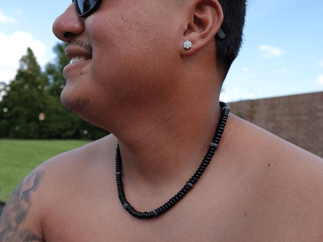 Close-up of a person's face, neck, and shoulder, wearing sunglasses, earrings, and a beaded necklace outdoors.