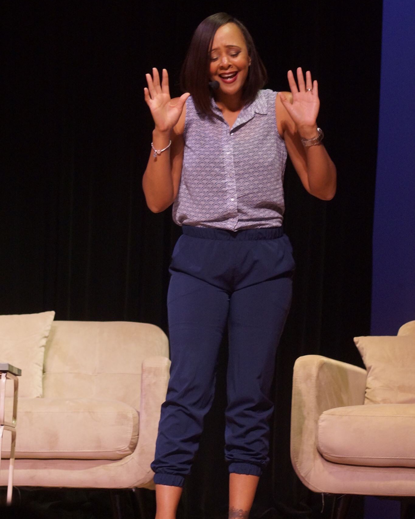 Woman standing on stage with arms raised, smiling, wearing a patterned shirt and dark pants, two beige sofas behind her.