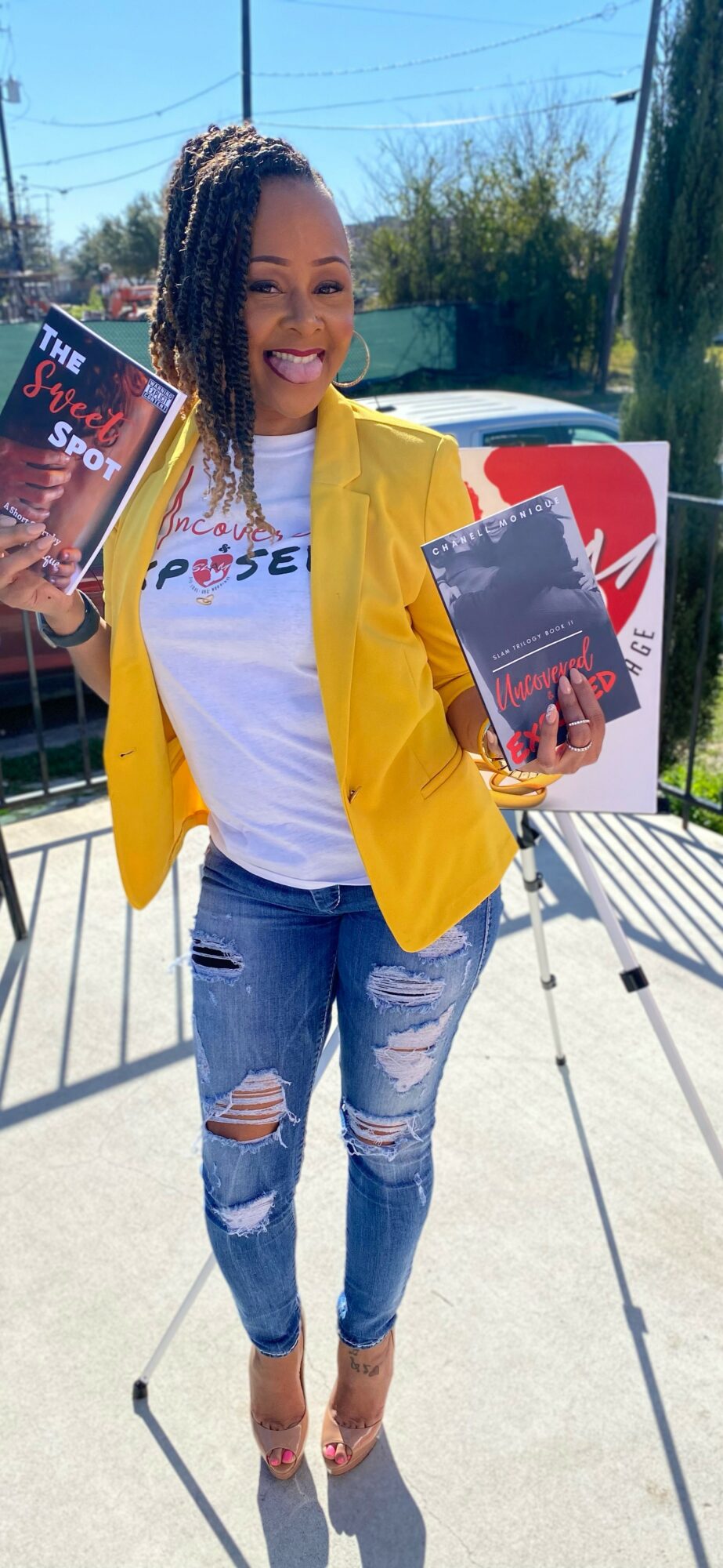 Woman with dreadlocks smiling, holding a book outdoors on a sunny day, wearing a yellow jacket and ripped jeans.