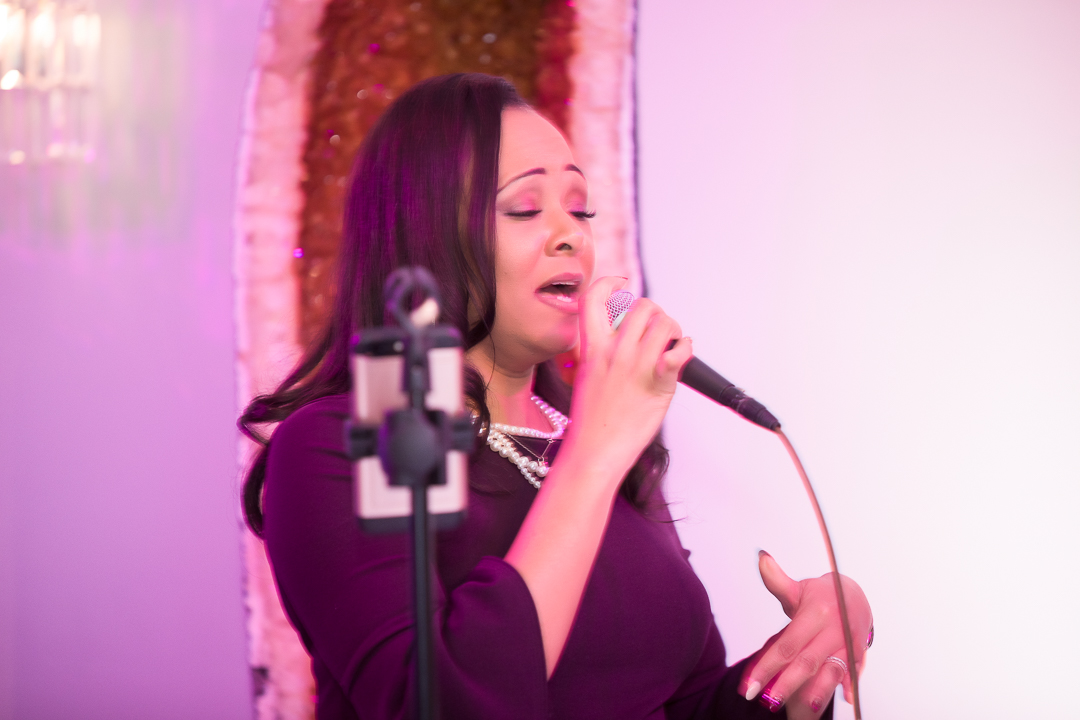 Woman singing into a microphone with a pink background, wearing a dark outfit and jewelry.