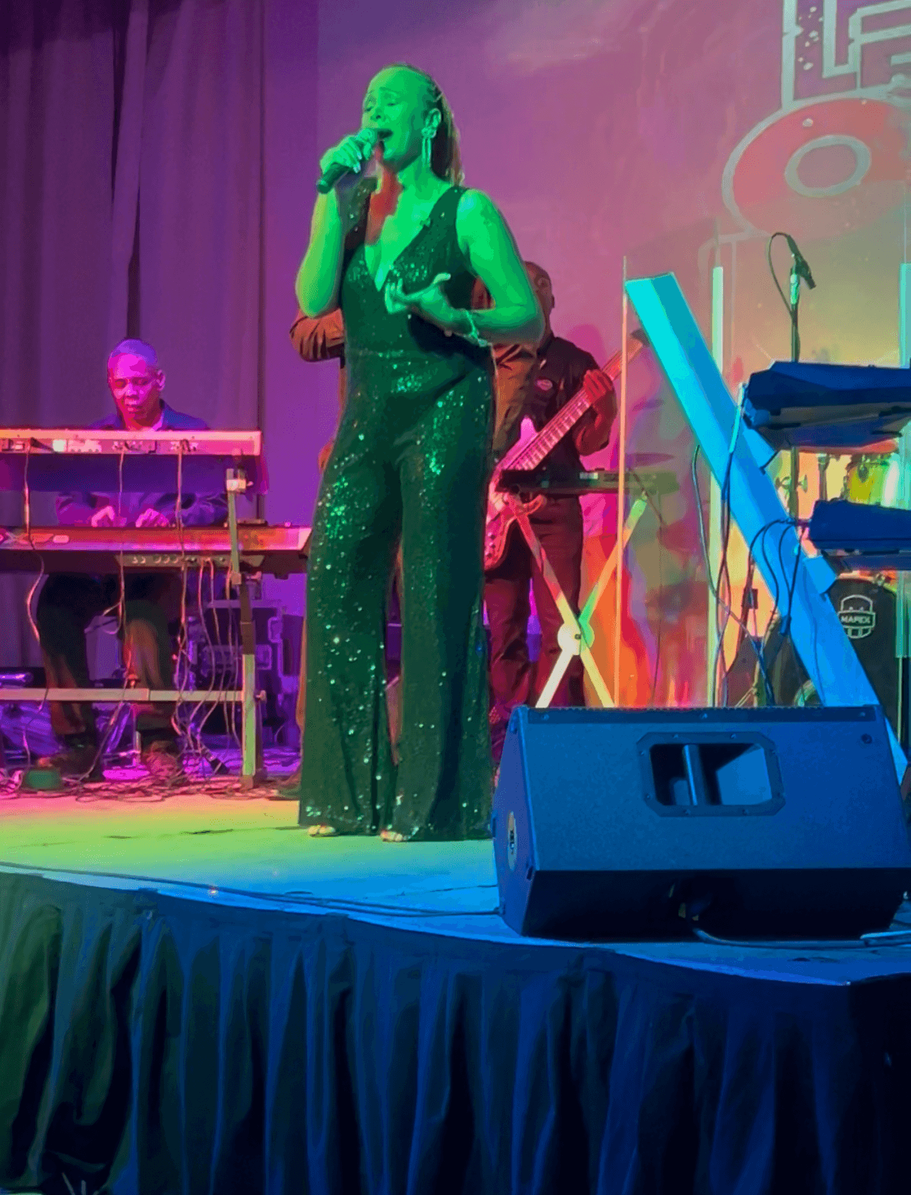 Singer in green dress performs on stage with band, colorful background, and musical equipment visible.