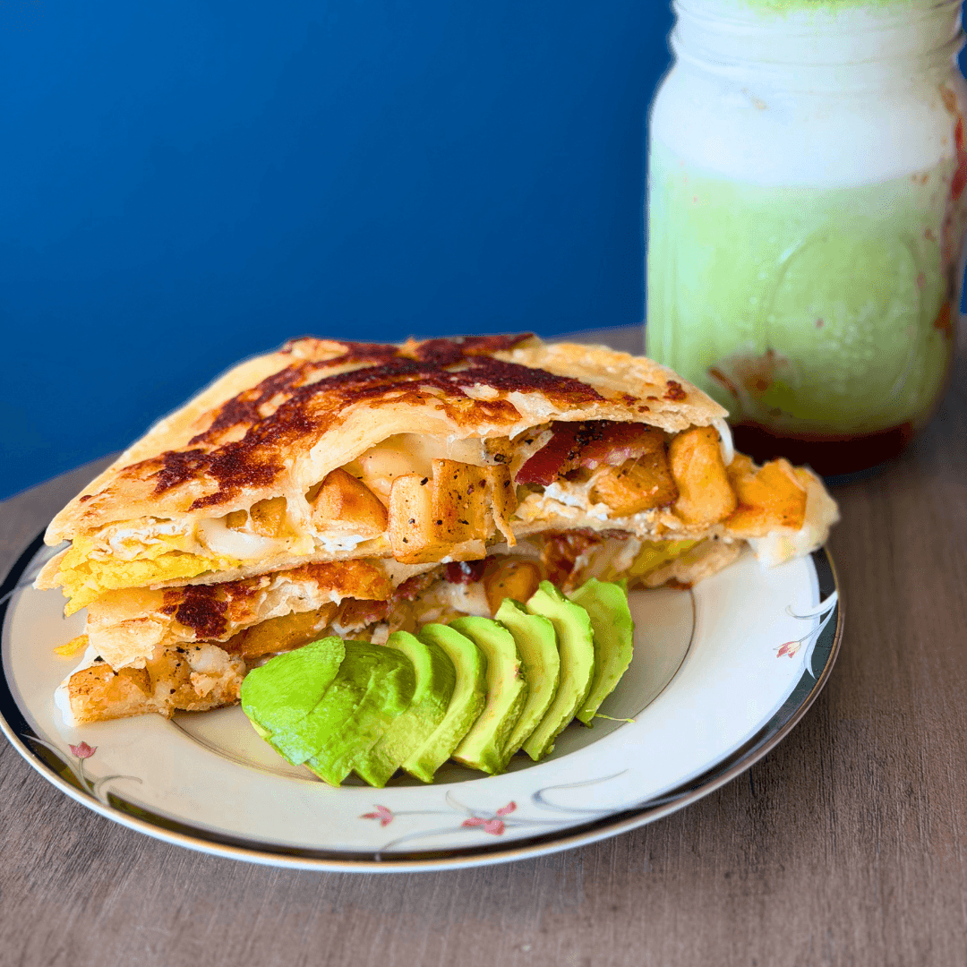 A slice of layered pie with vegetables and cheese, served with sliced avocado on a decorative plate, against a blue background.