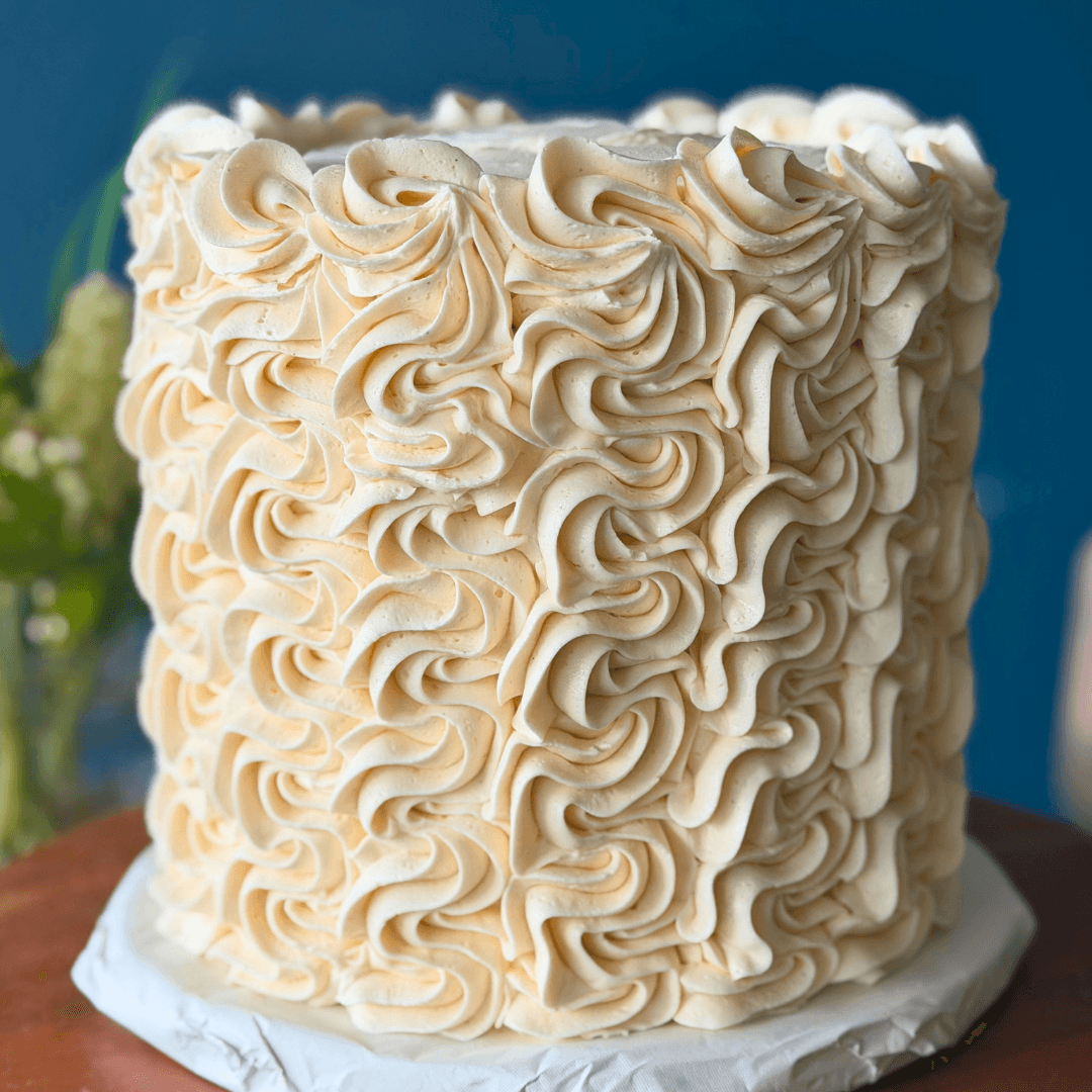 A tall, round cake decorated with cream-colored ruffled frosting, placed on a white base, with a blurred background.