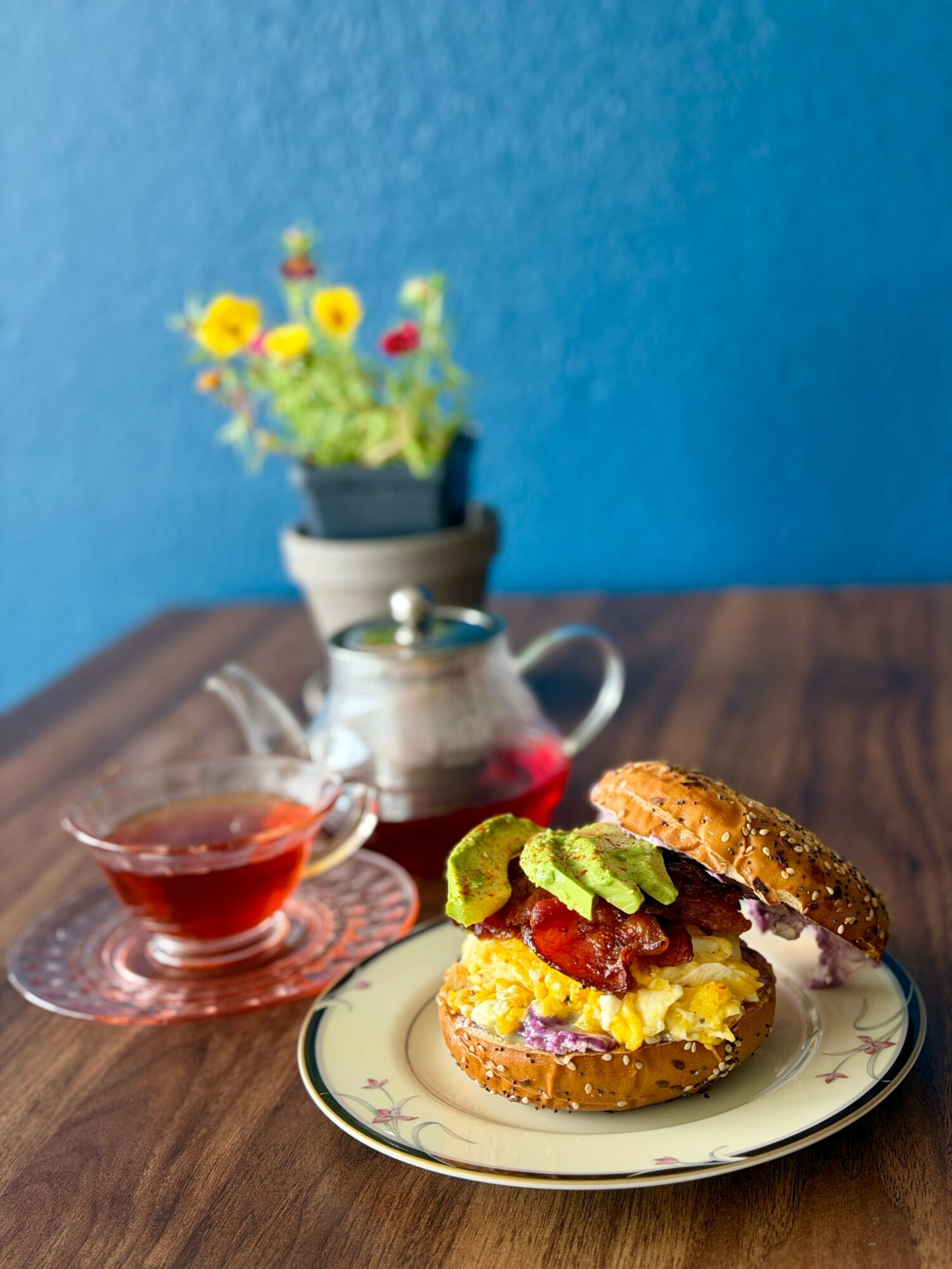 A breakfast sandwich with bacon, lettuce, and egg on a plate, with a small pitcher of syrup and a flower pot in the background.