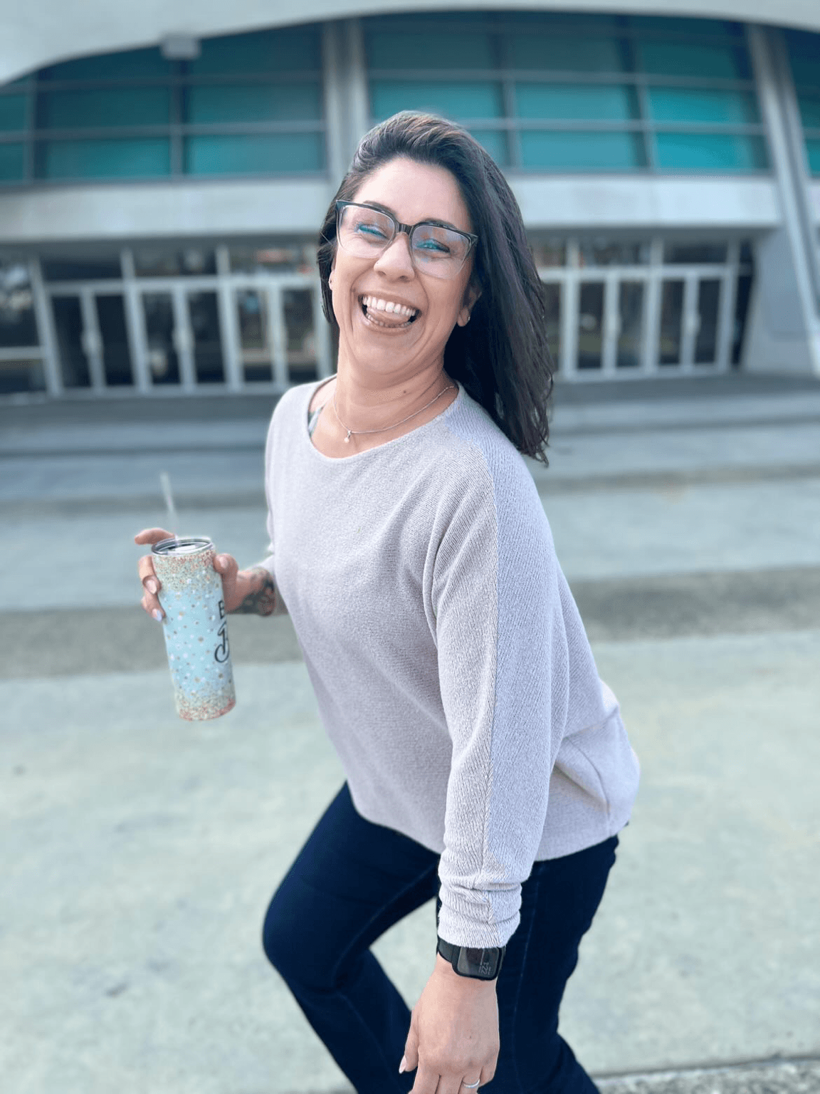 Woman with glasses smiling, holding a drink, standing outdoors in front of a modern building.