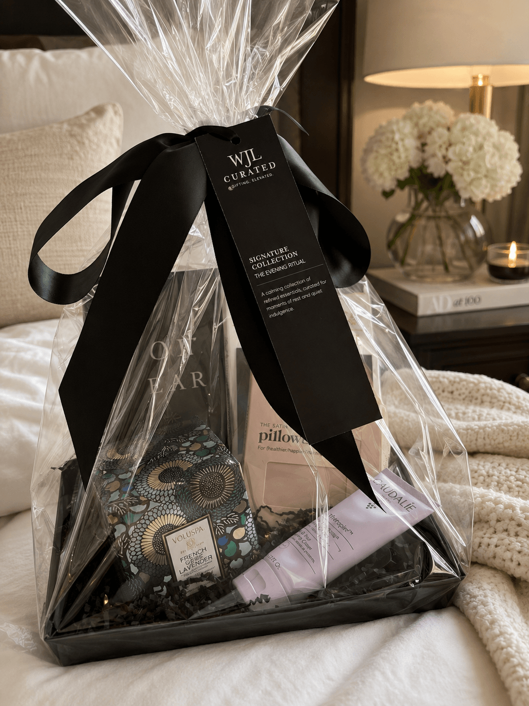 Gift wrapped in clear plastic with a black ribbon and tag, placed on a bed with a nightstand and flowers in background.
