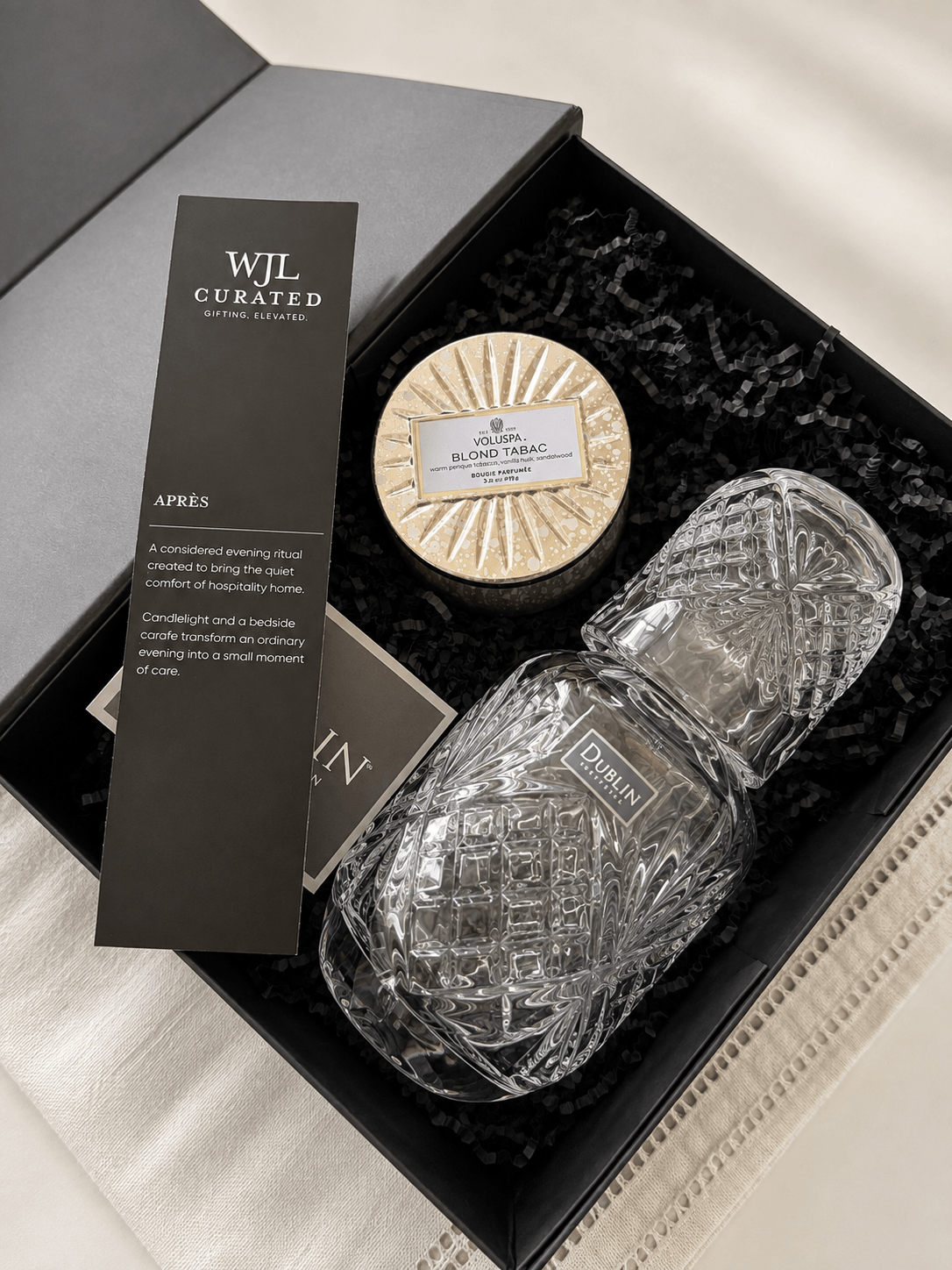 Open black box with a round soap and a clear textured bottle inside, black shredded paper lining the box, and a black card.