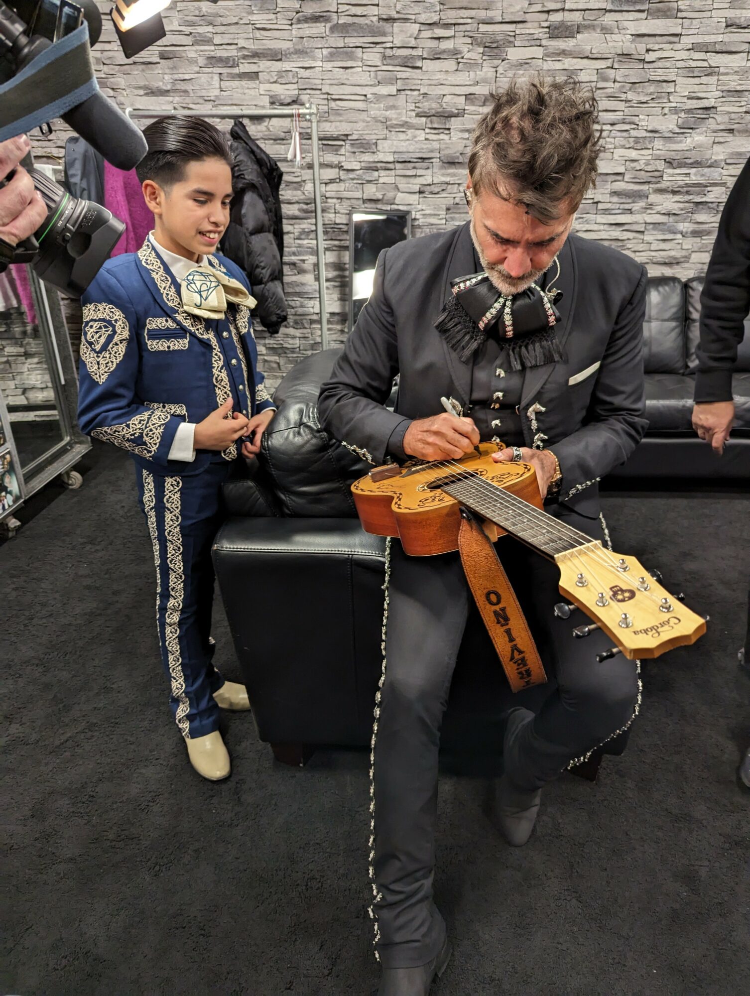 Man sitting on a bench signing a guitar, young girl standing nearby watching, in a room with a stone wall background.