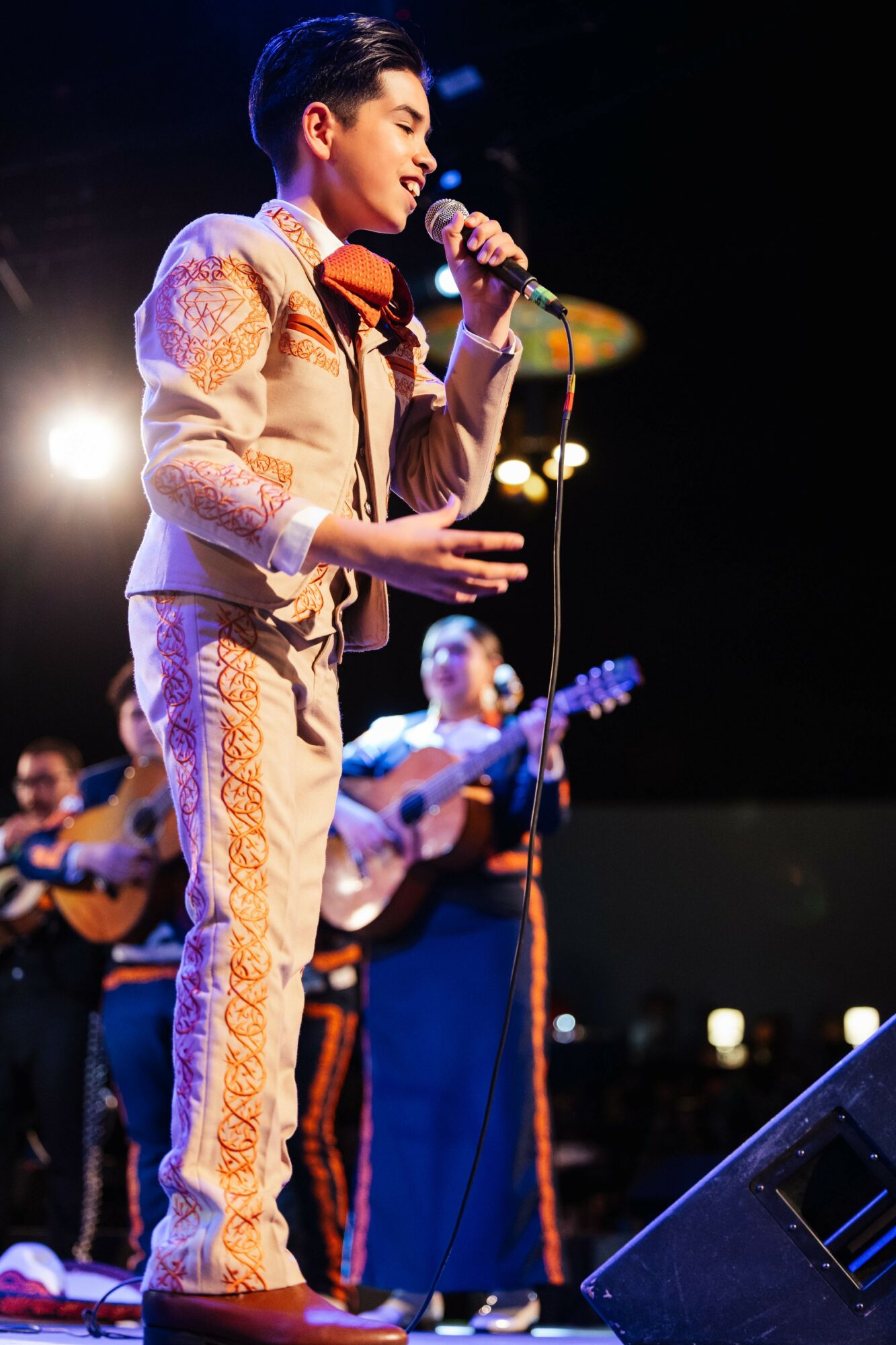 Young man singing into a microphone on stage with guitar players in the background, stage lights shining brightly.