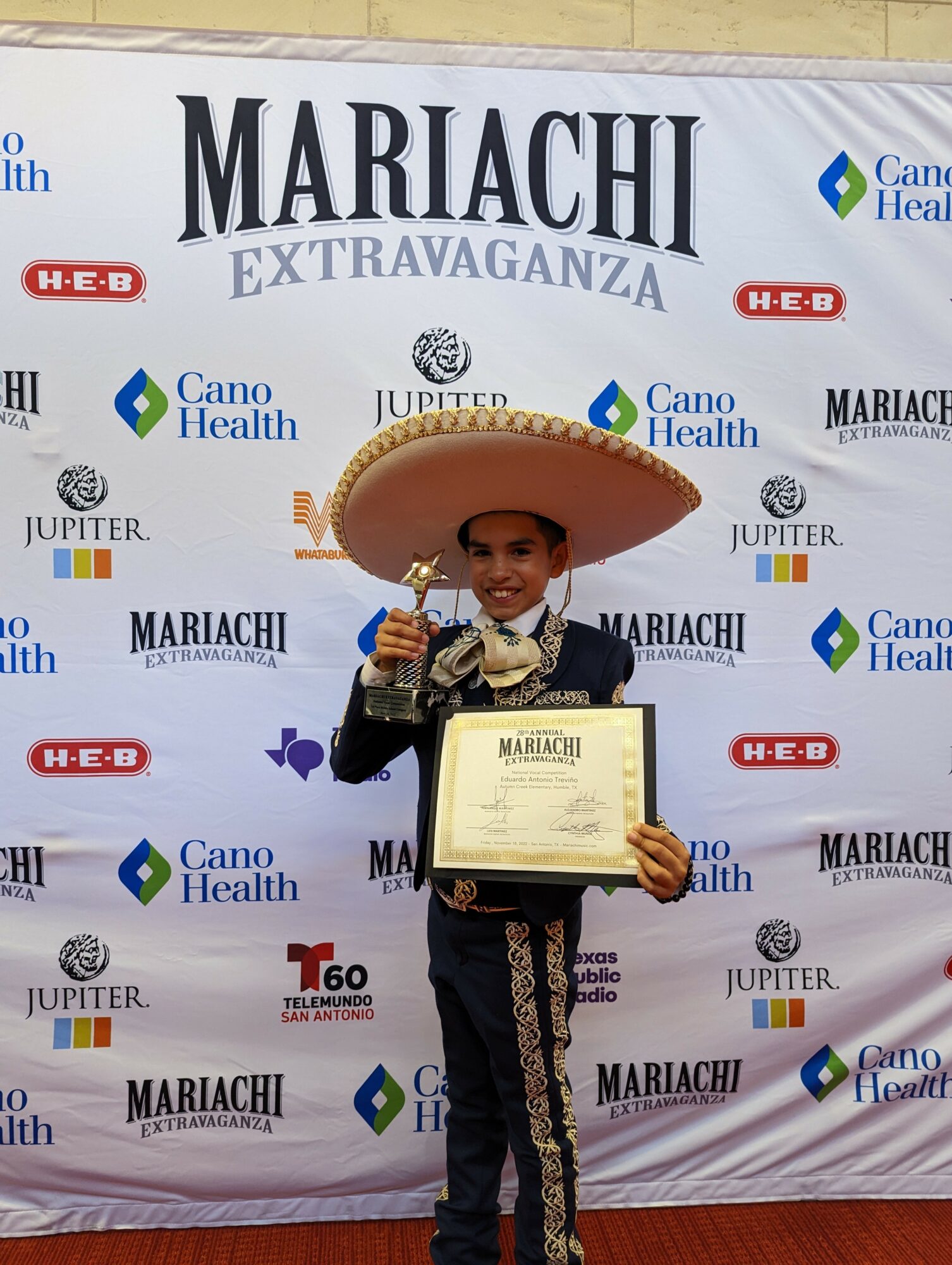 Child in traditional Mexican attire holding a trophy and certificate, standing in front of a step-and-repeat banner with event logos.