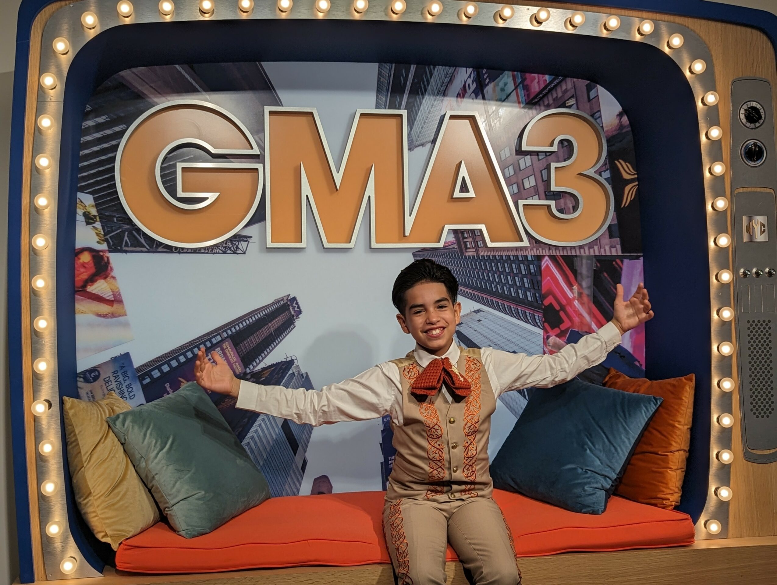 Young boy in beige suit with bow tie, smiling with arms outstretched, sitting on a colorful stage with pillows and a GMA3 sign.